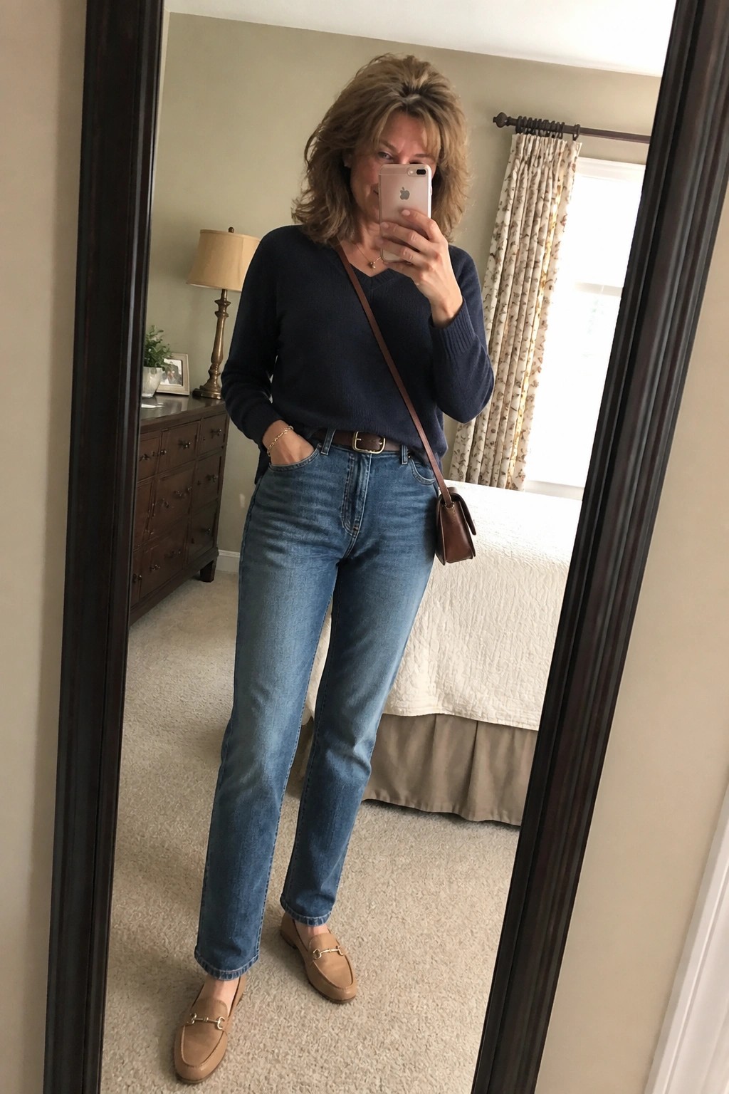 Woman in navy sweater and jeans taking mirror selfie in bedroom