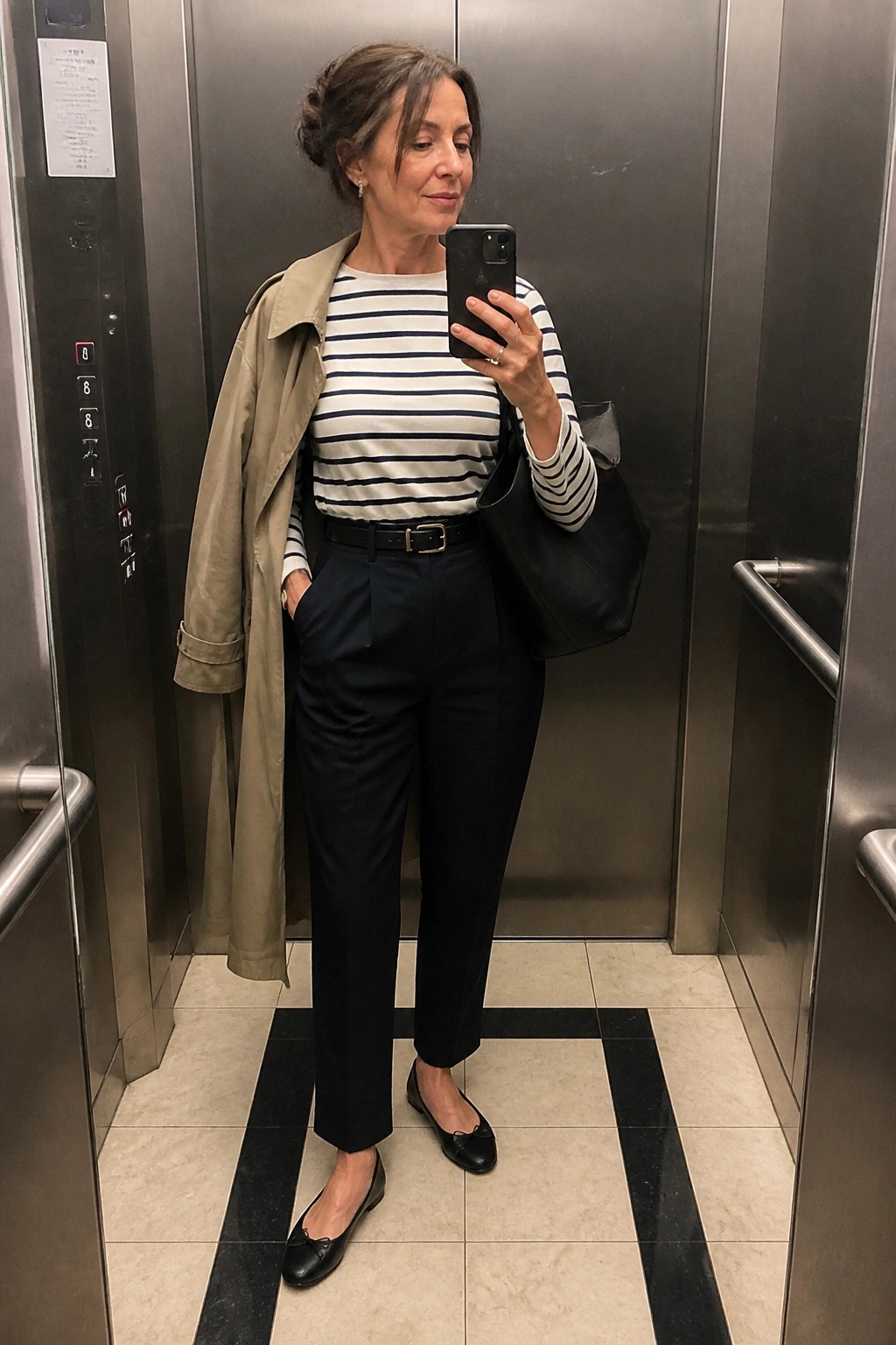 Woman in striped shirt and navy pants taking selfie in elevator with black bag