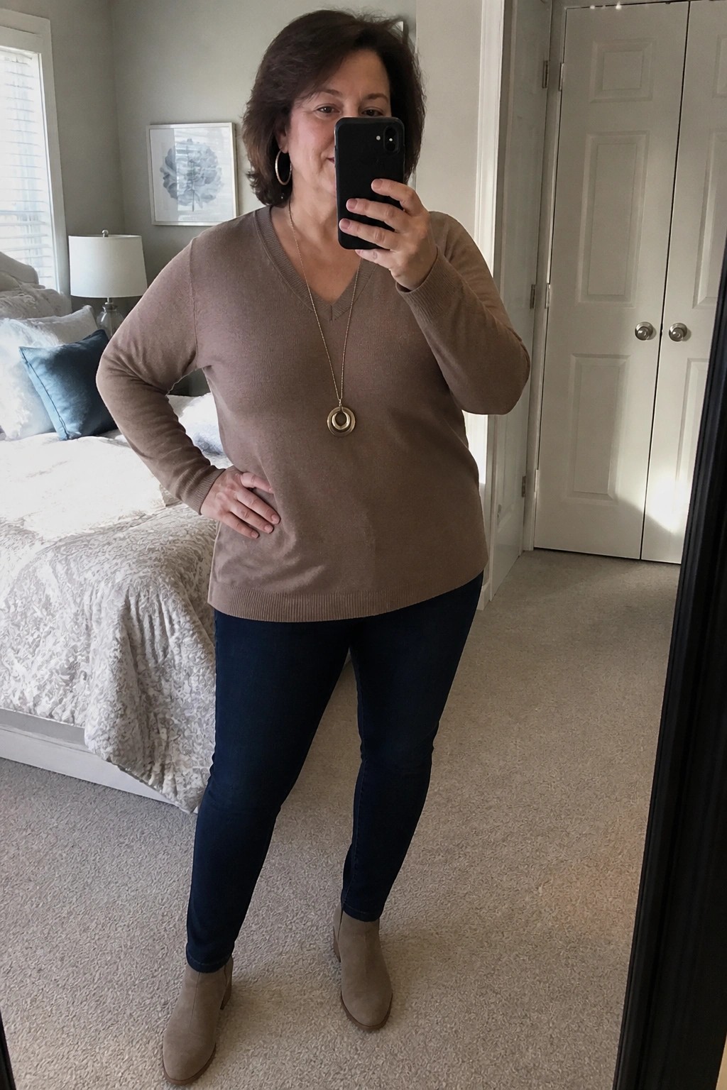Woman over 50 wearing a taupe V-neck sweater tucked slightly at the front with dark blue jeans and taupe suede ankle boots, accessorized with a long gold necklace featuring a circular pendant, taking a mirror selfie in a bedroom setting.