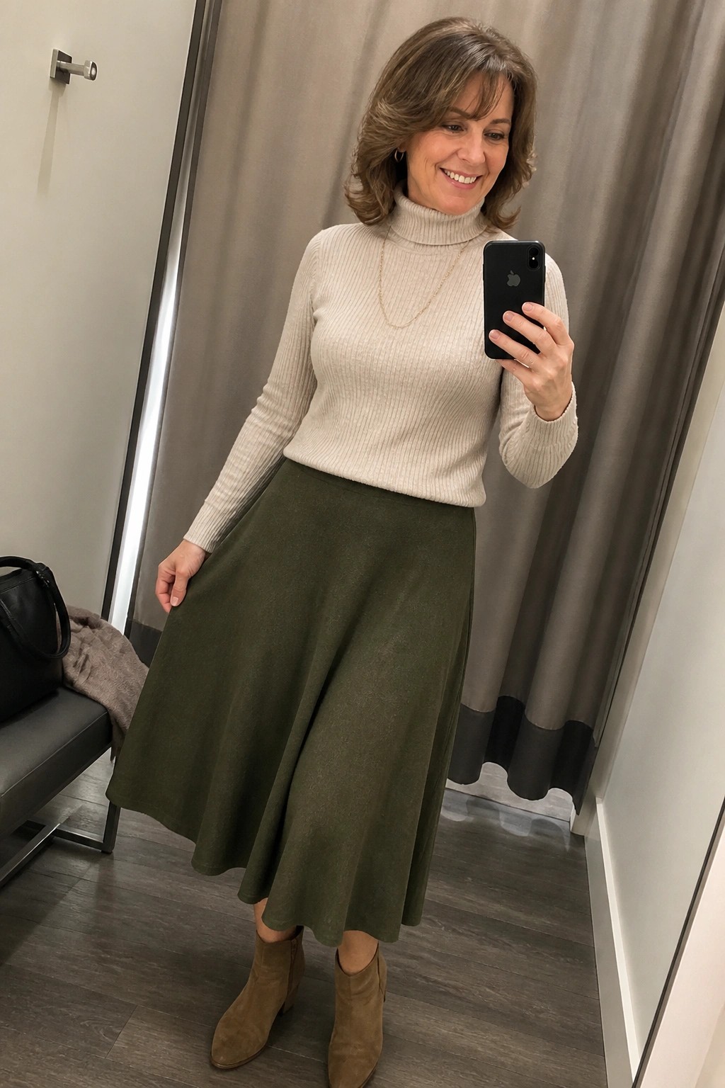 Woman wearing a beige ribbed turtleneck sweater with a long gold necklace, an olive green A-line midi skirt, and taupe suede ankle boots while holding a phone for a mirror selfie.