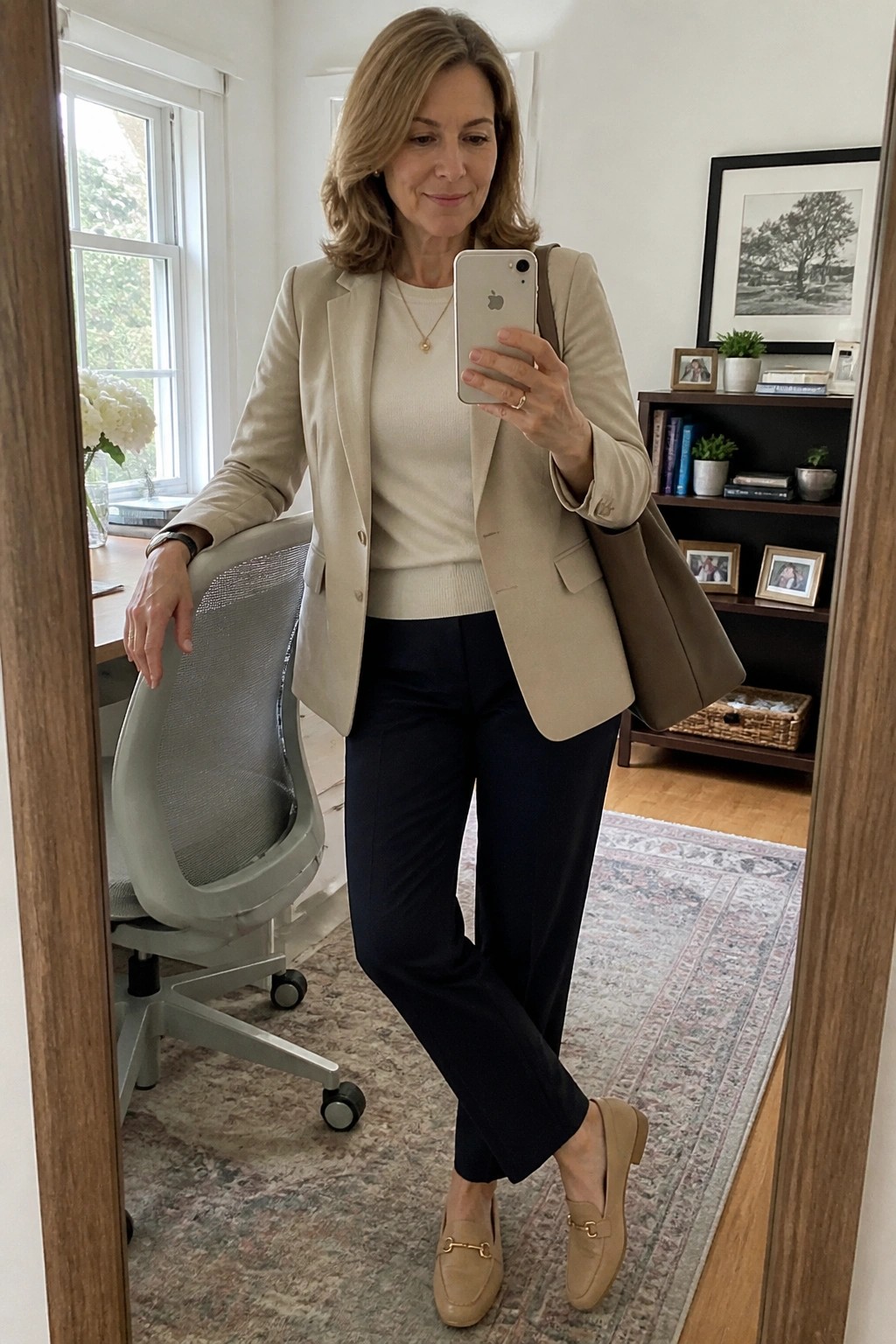 Woman over 50 in a beige blazer layered over a cream sweater with navy pants and beige loafers holding a phone for a mirror selfie.