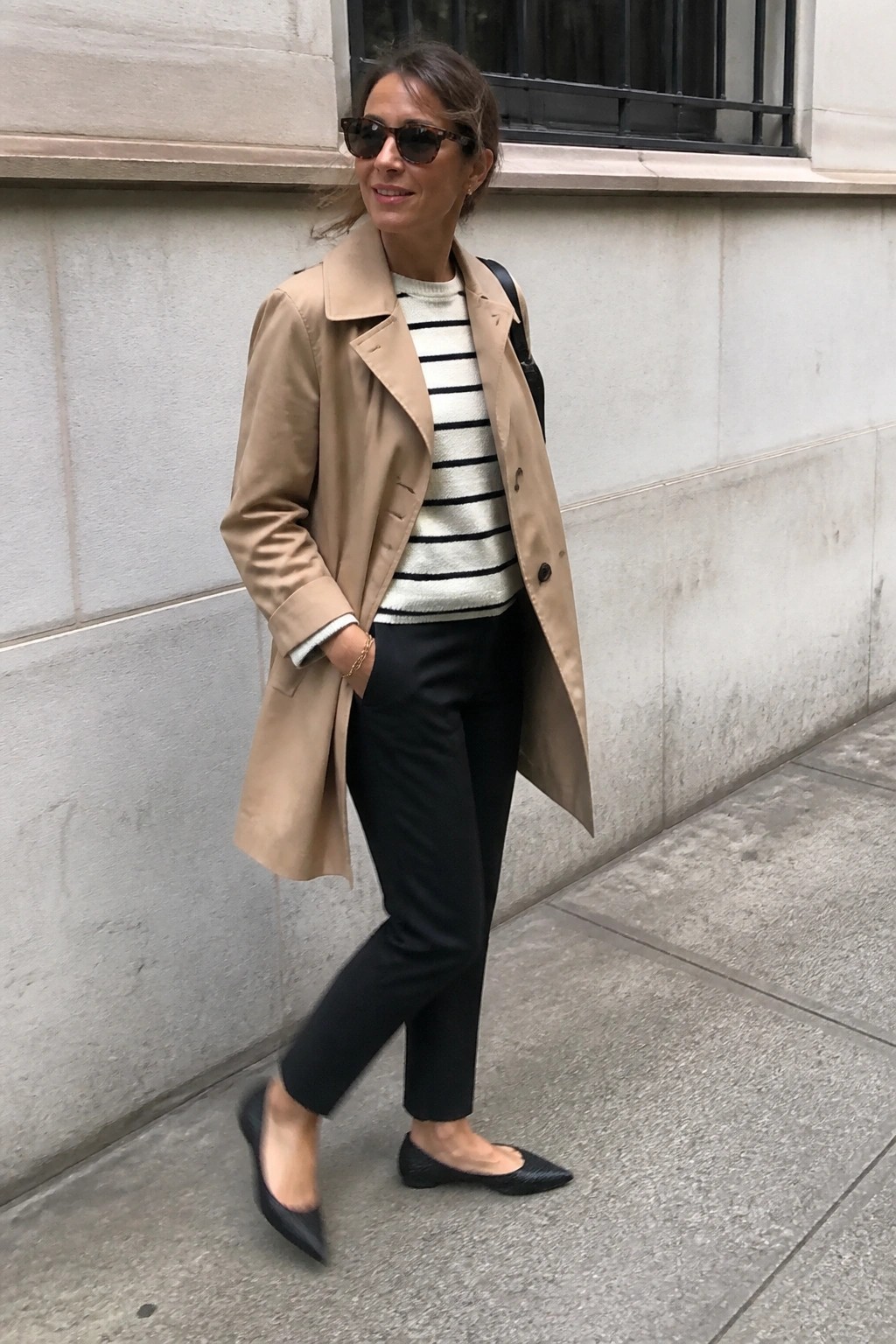 Woman in beige trench coat, striped sweater, black pants, and flats by stone wall
