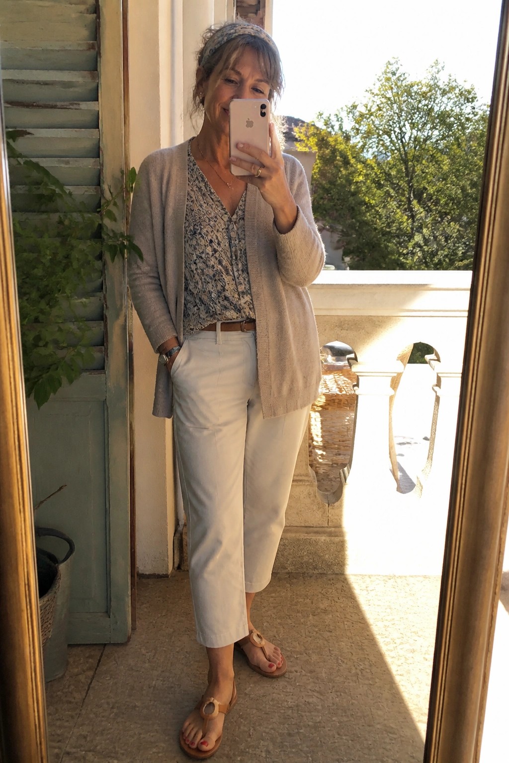 Woman in beige cardigan, floral top, and white pants taking balcony selfie