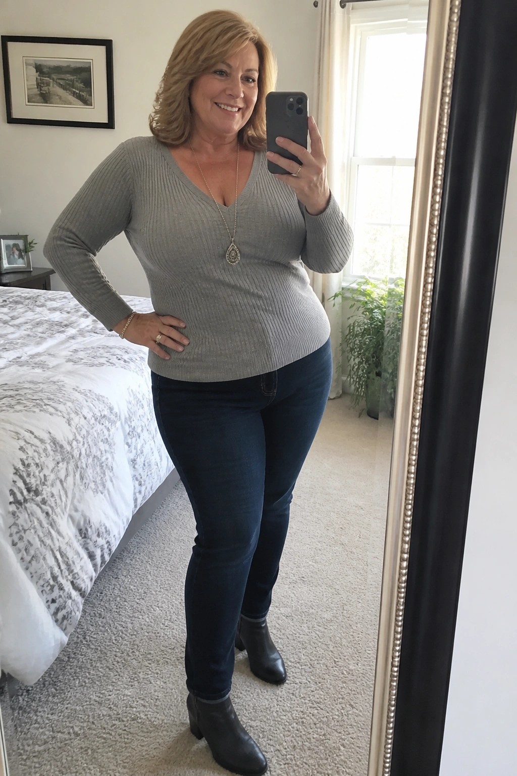 Blonde woman in gray sweater and jeans taking mirror selfie in bedroom.