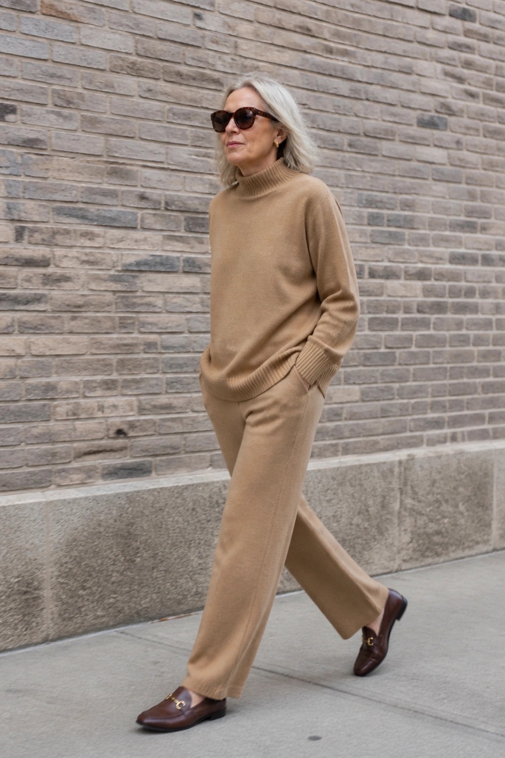 Gray-haired woman in tan turtleneck sweater and wide pants by brick wall.