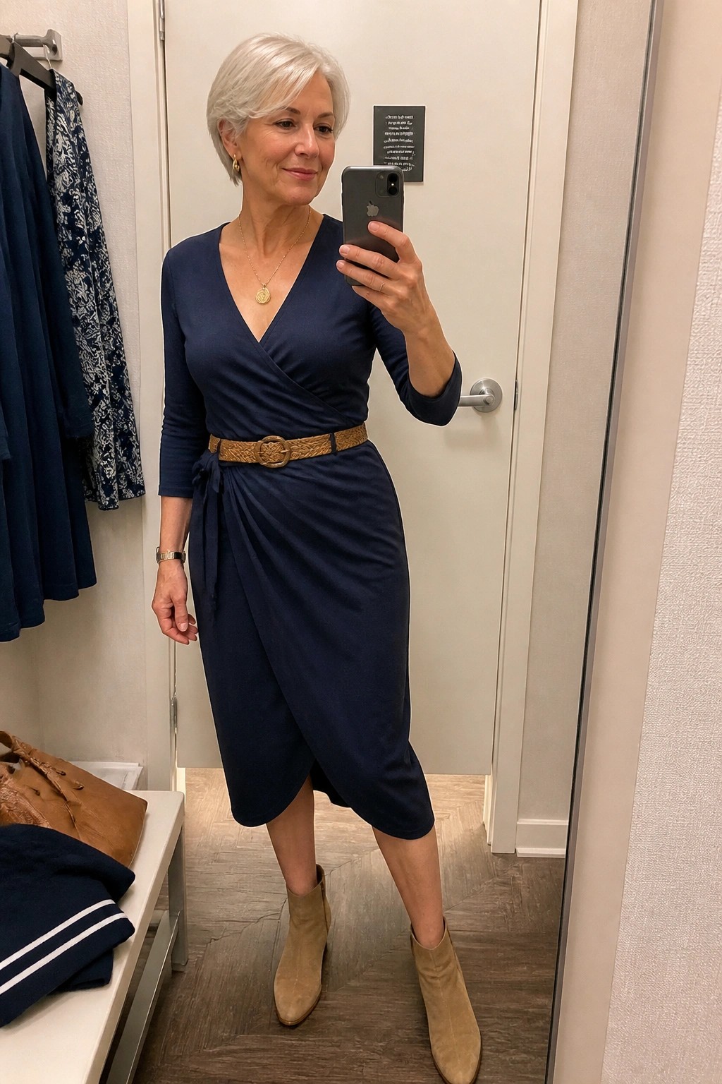 Woman in navy wrap dress with tan belt and suede boots taking mirror selfie
