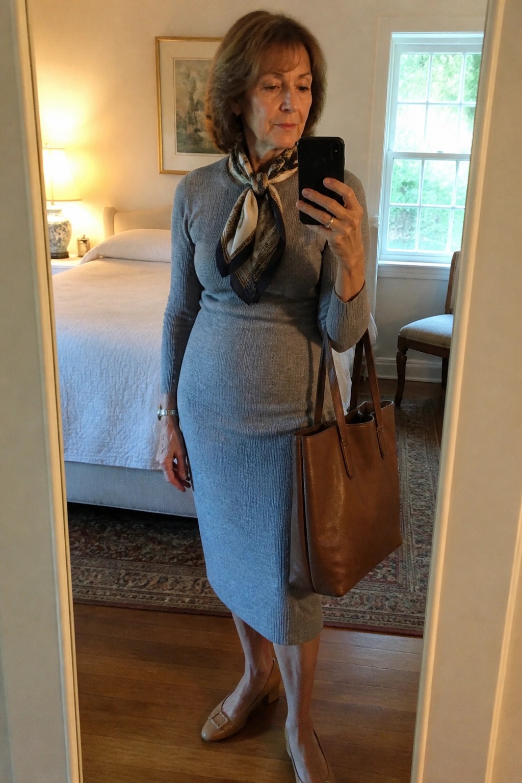 Older woman in blue knit dress with scarf holding tote for mirror selfie in bedroom