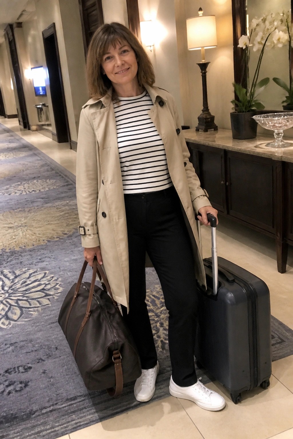 Woman in beige trench coat with striped top holding duffel bag and suitcase in hallway