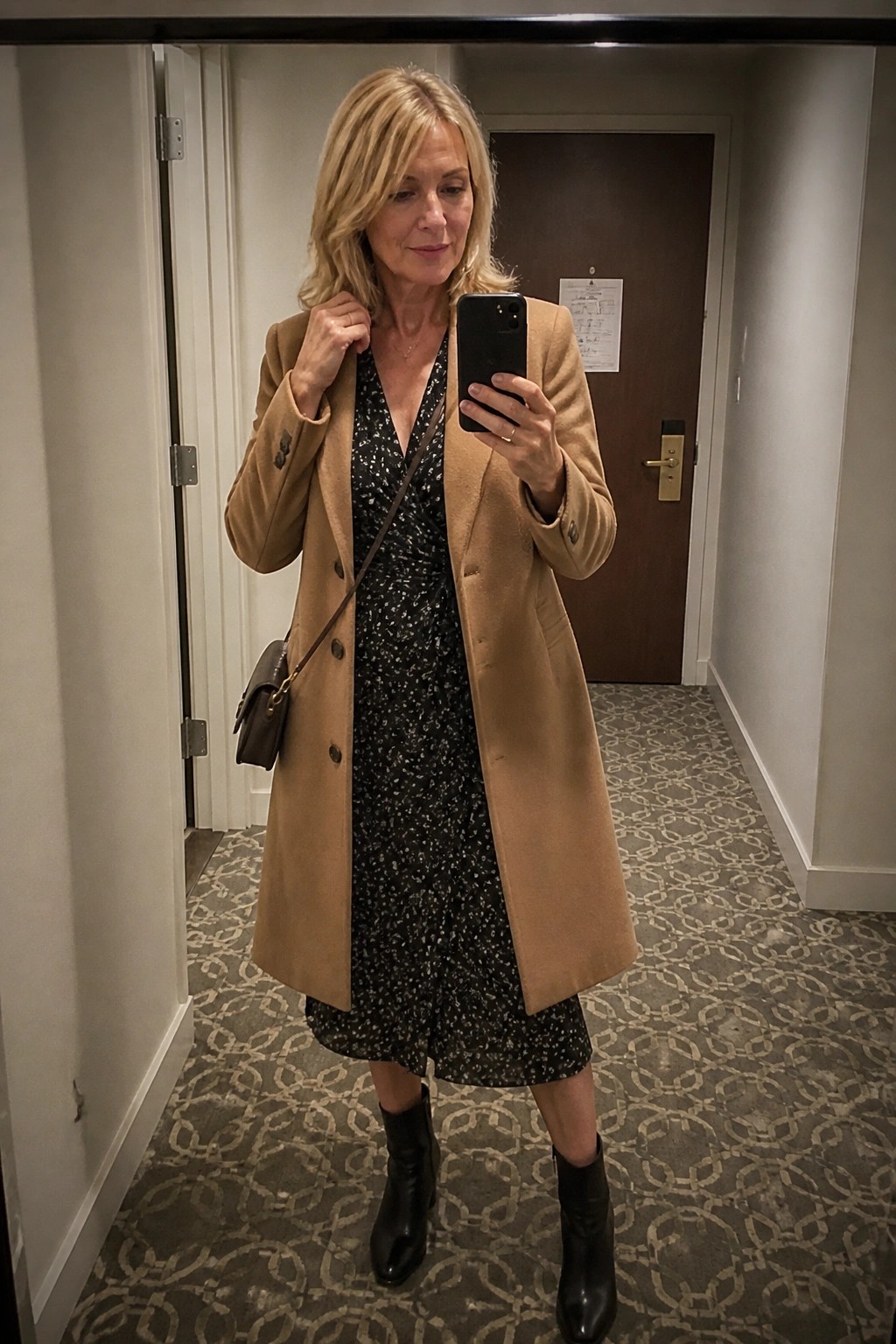 Blonde woman in tan coat over black floral dress taking mirror selfie in hallway