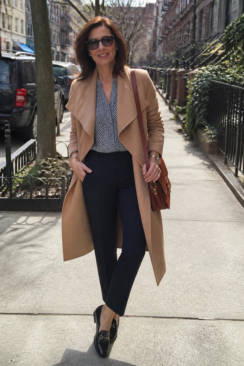 Woman in tan coat over patterned blouse and black pants on city sidewalk
