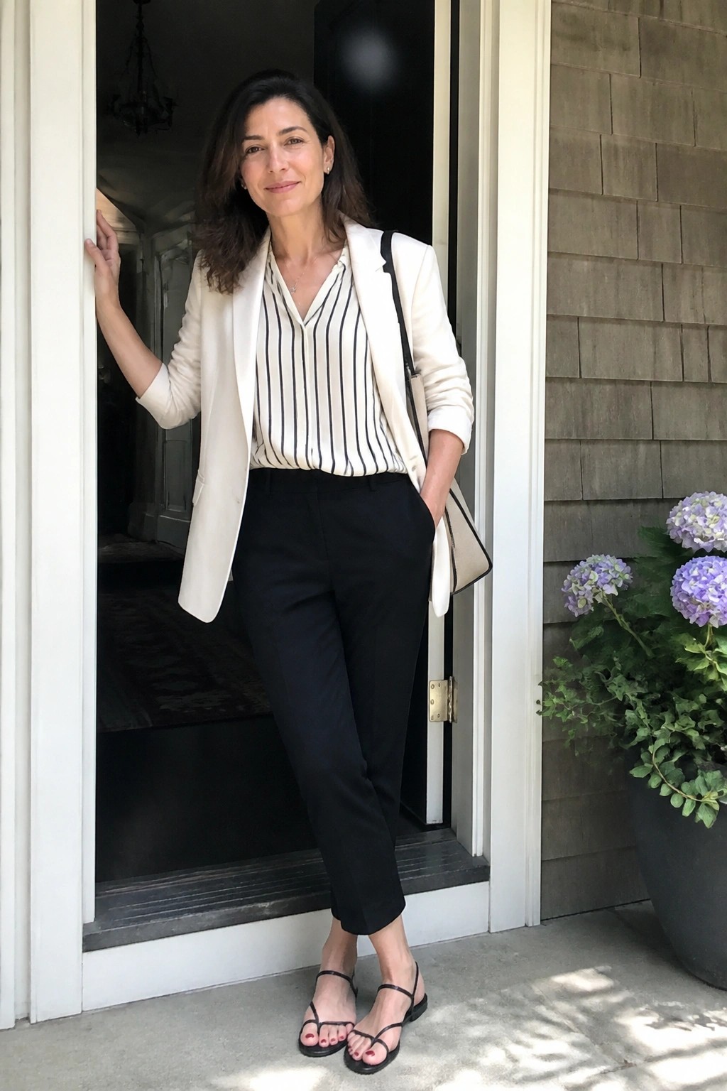 Woman in white blazer and striped blouse stands in doorway with black pants and handbag