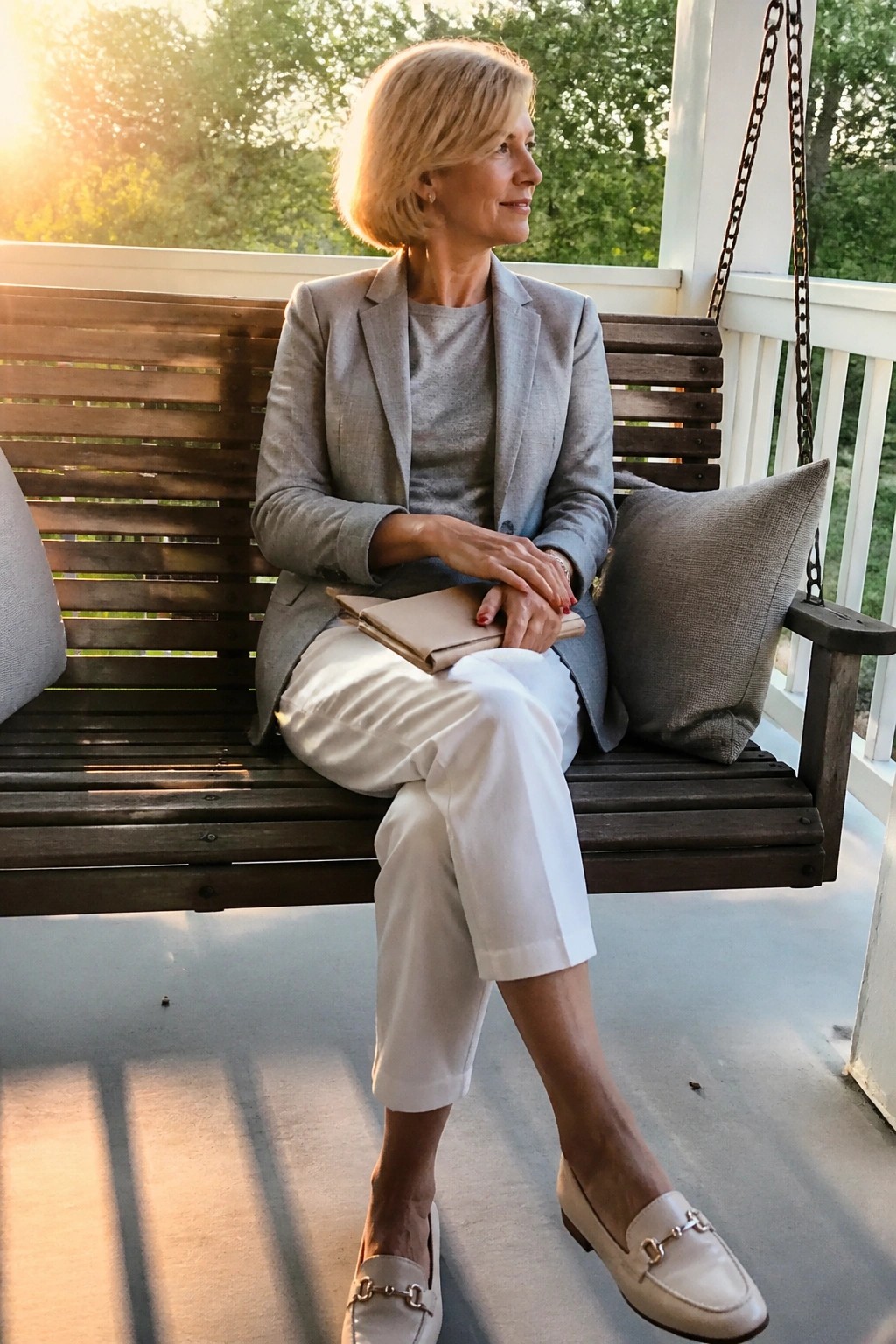Woman in gray blazer and white pants seated on wooden porch swing at sunset