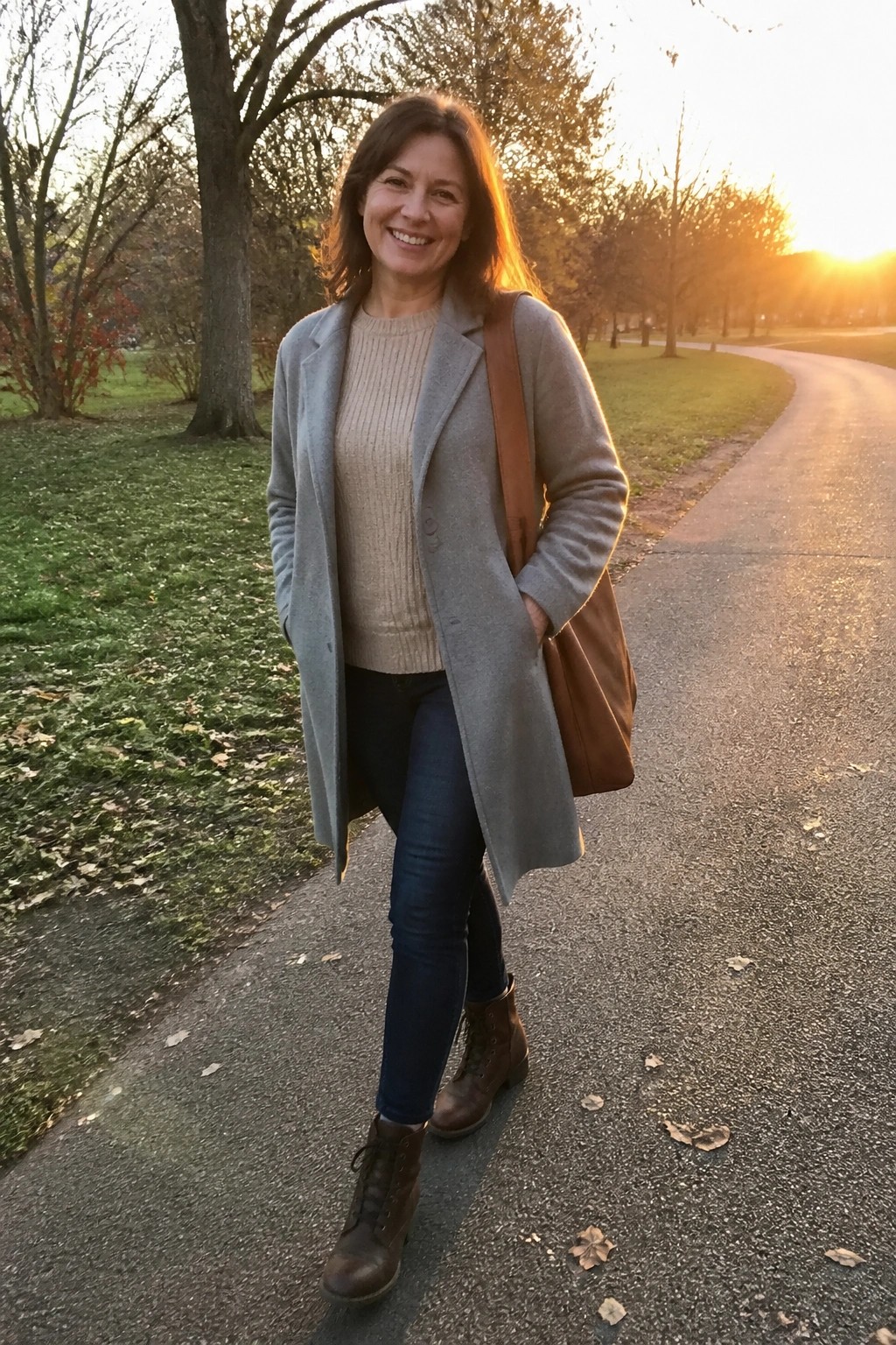 Smiling woman in gray coat over beige sweater on autumn park path at sunset