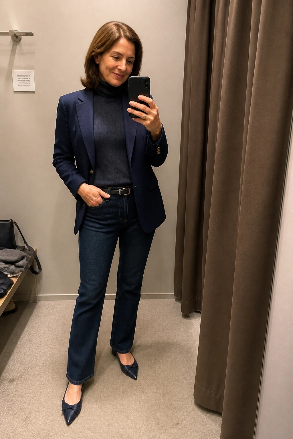 Woman in navy blazer, turtleneck, and jeans taking a mirror selfie in fitting room