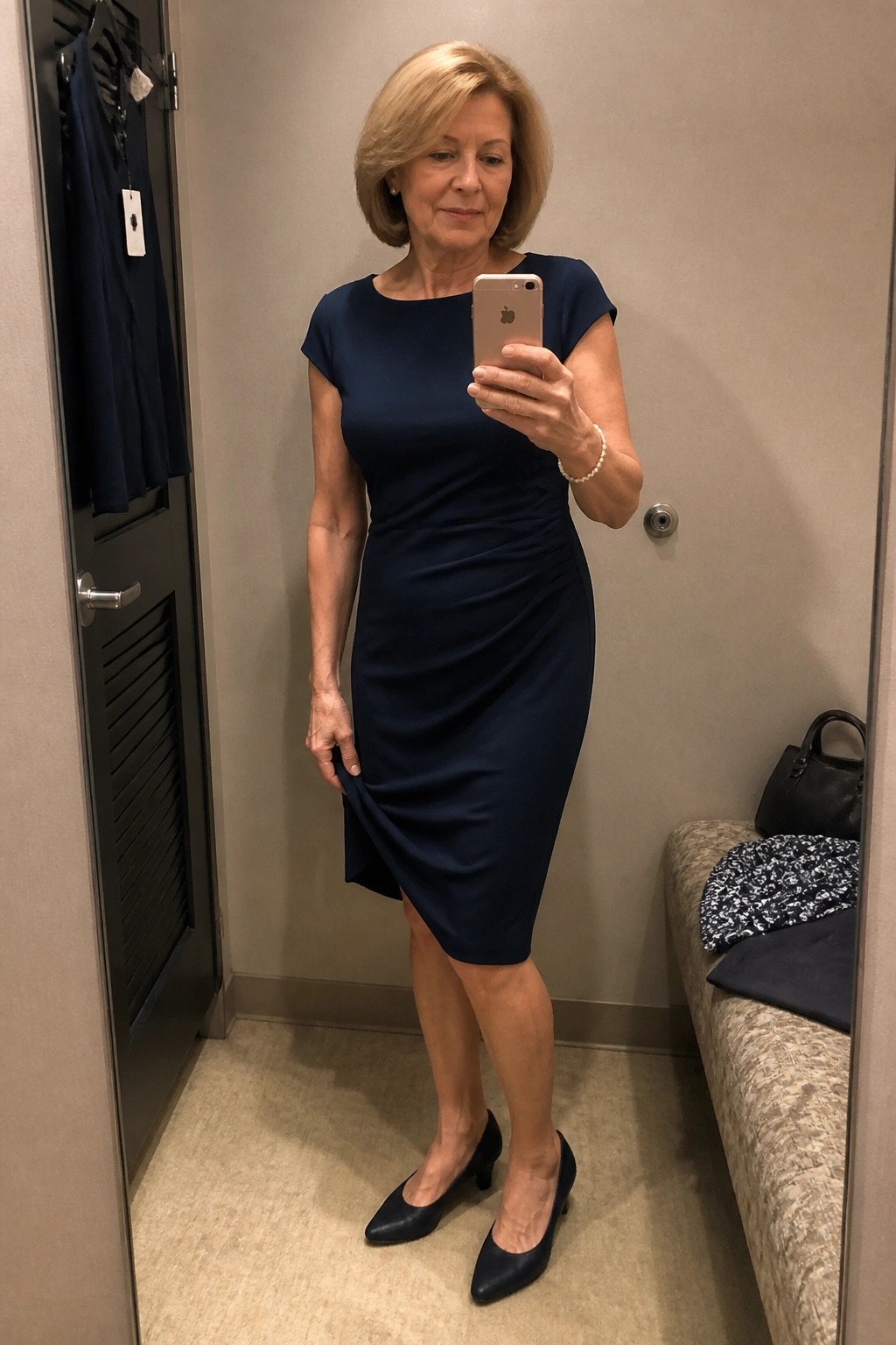 Woman in navy blue dress taking mirror selfie in fitting room holding iPhone