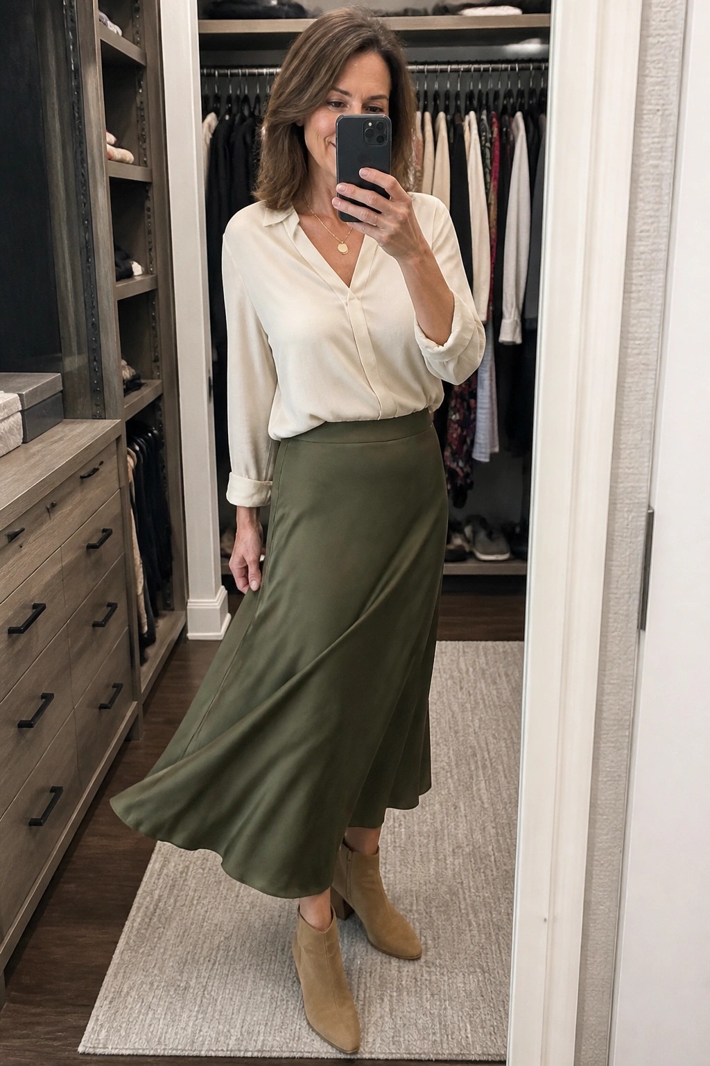 Woman in cream blouse and olive skirt taking mirror selfie in closet