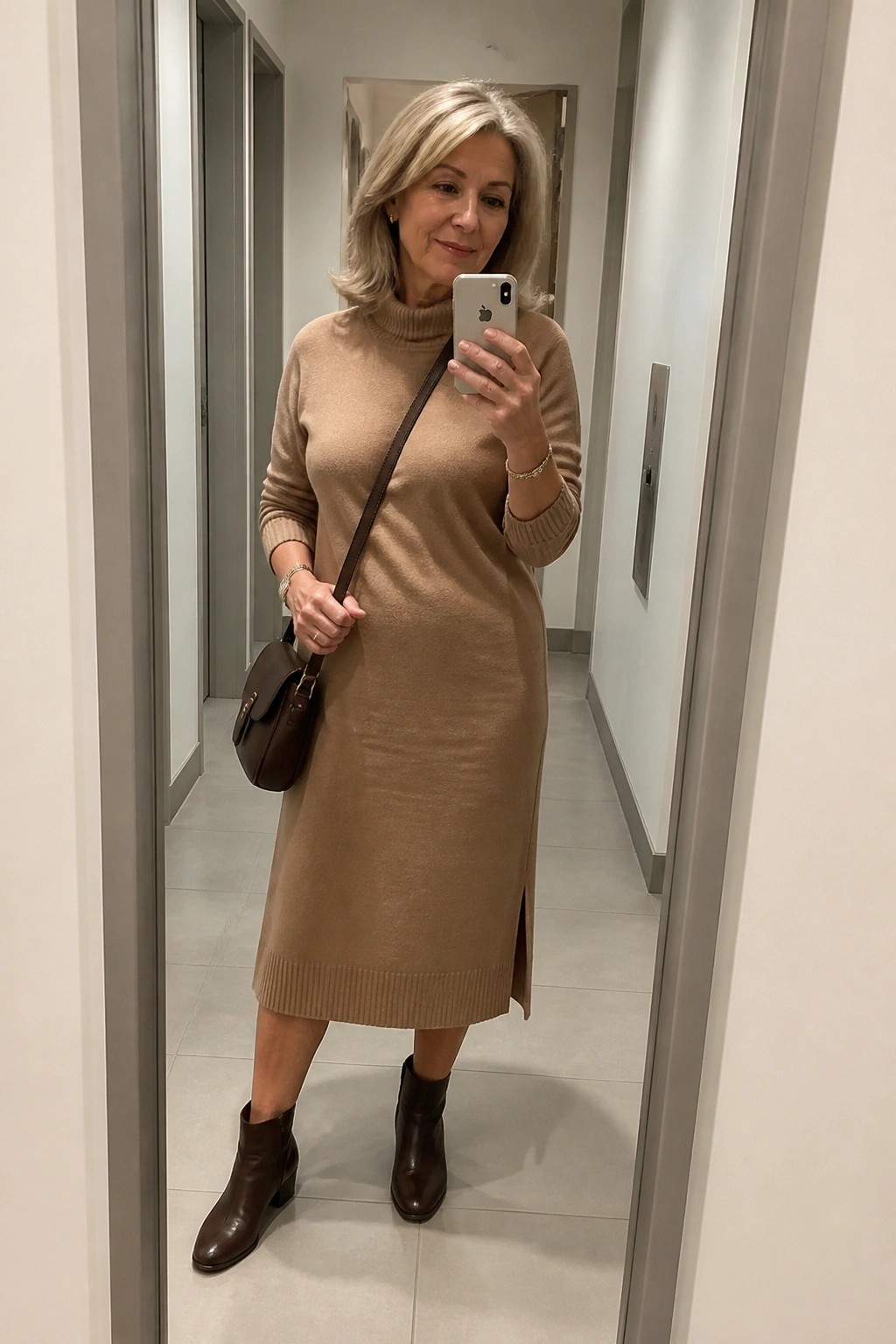 Woman in beige turtleneck sweater dress and brown boots taking mirror selfie in hallway