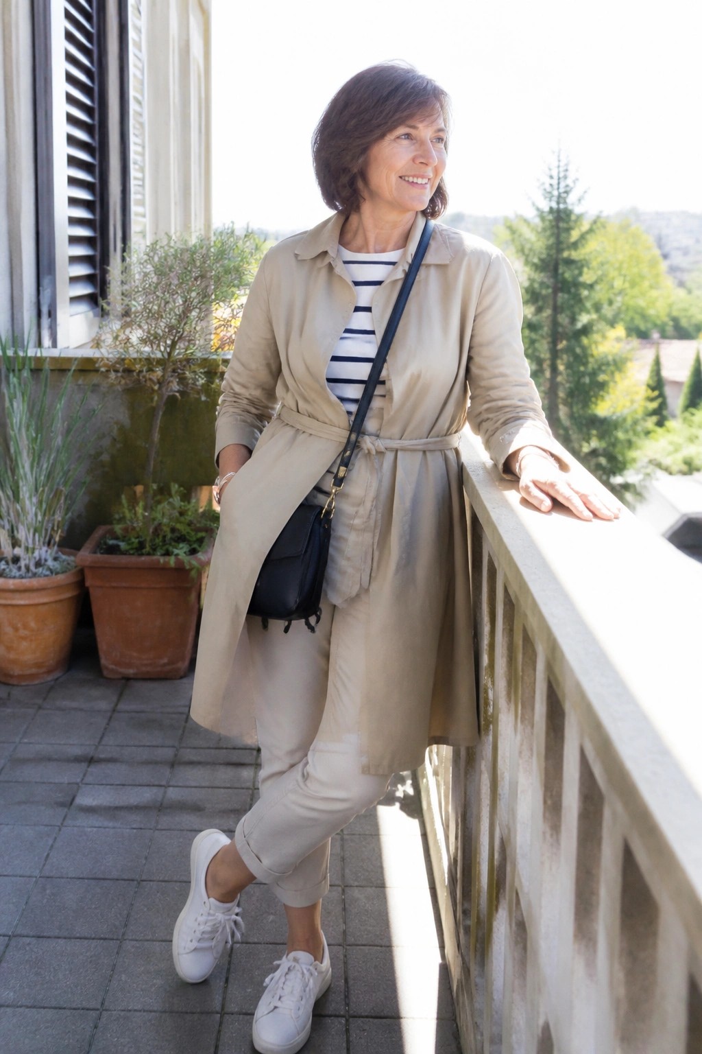 Woman in beige trench coat and striped shirt leaning on sunny balcony railing