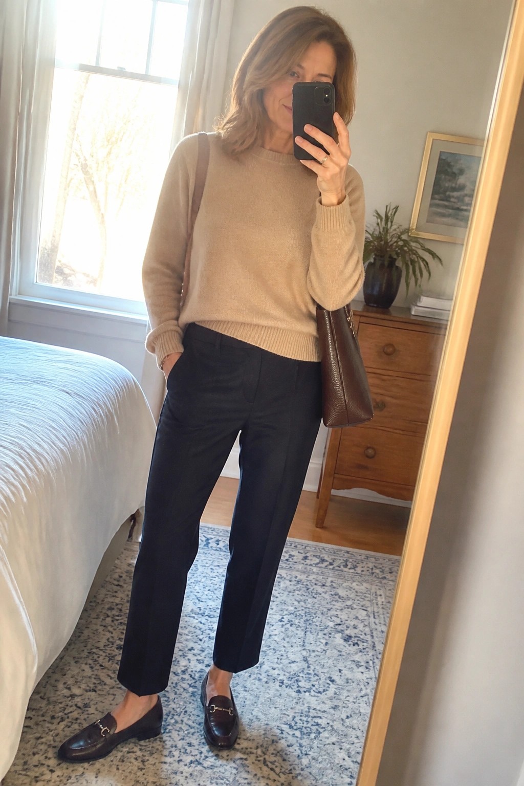 Woman in beige sweater and black pants taking bedroom mirror selfie with phone