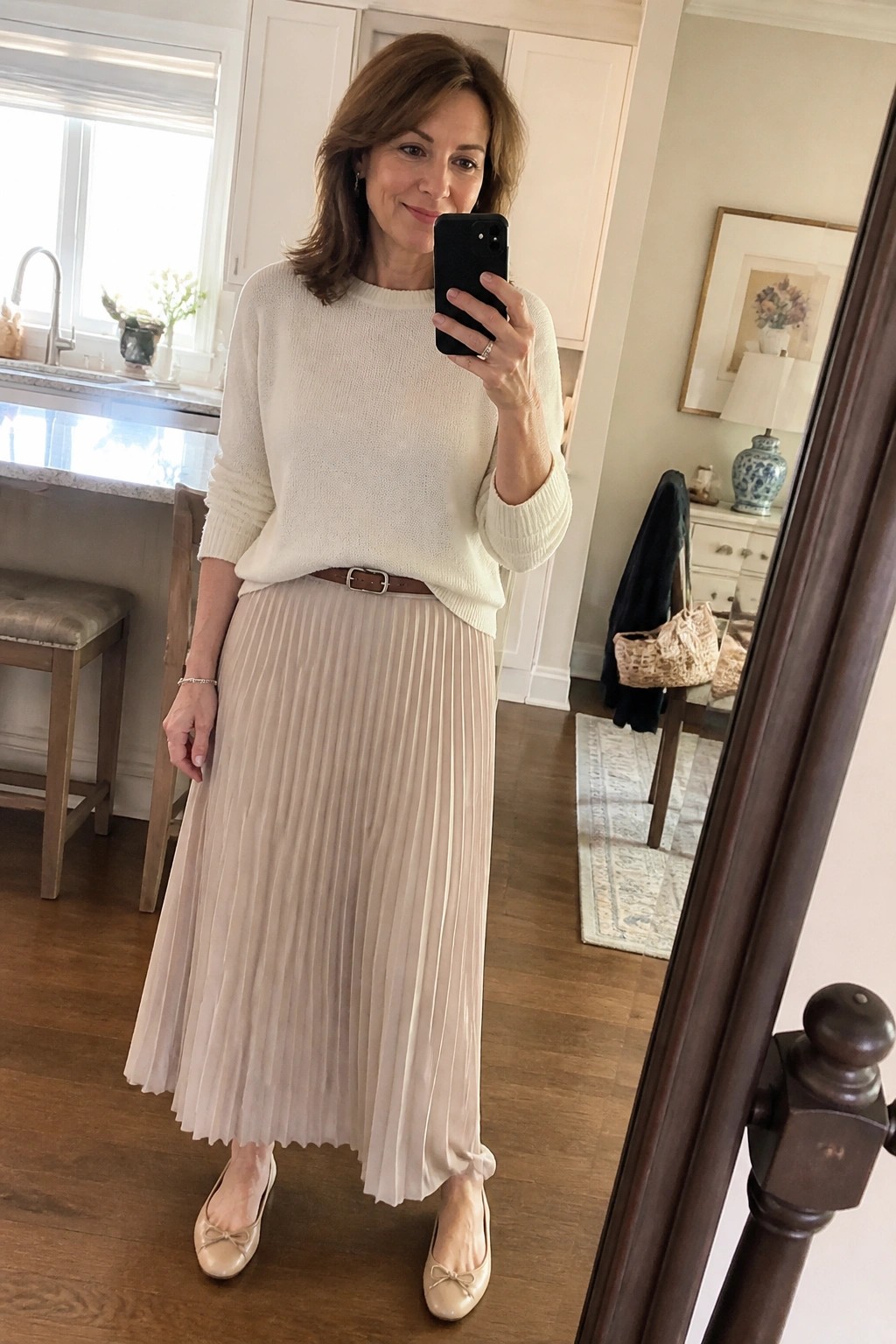 Woman in cream sweater and beige pleated skirt taking mirror selfie in home