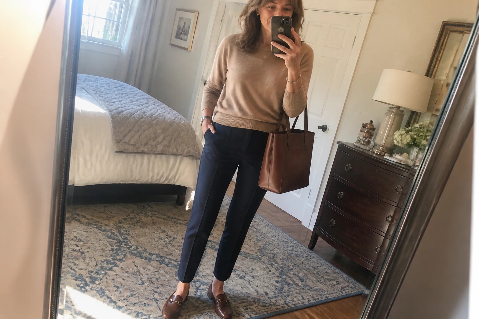 Woman in beige sweater and black pants taking bedroom mirror selfie with phone