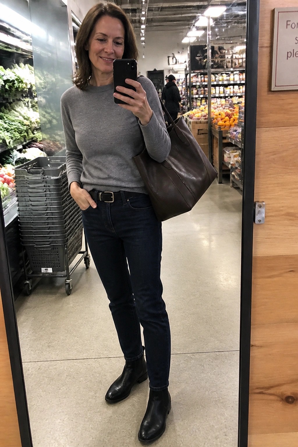 Woman in gray sweater and jeans takes mirror selfie with brown tote in grocery store