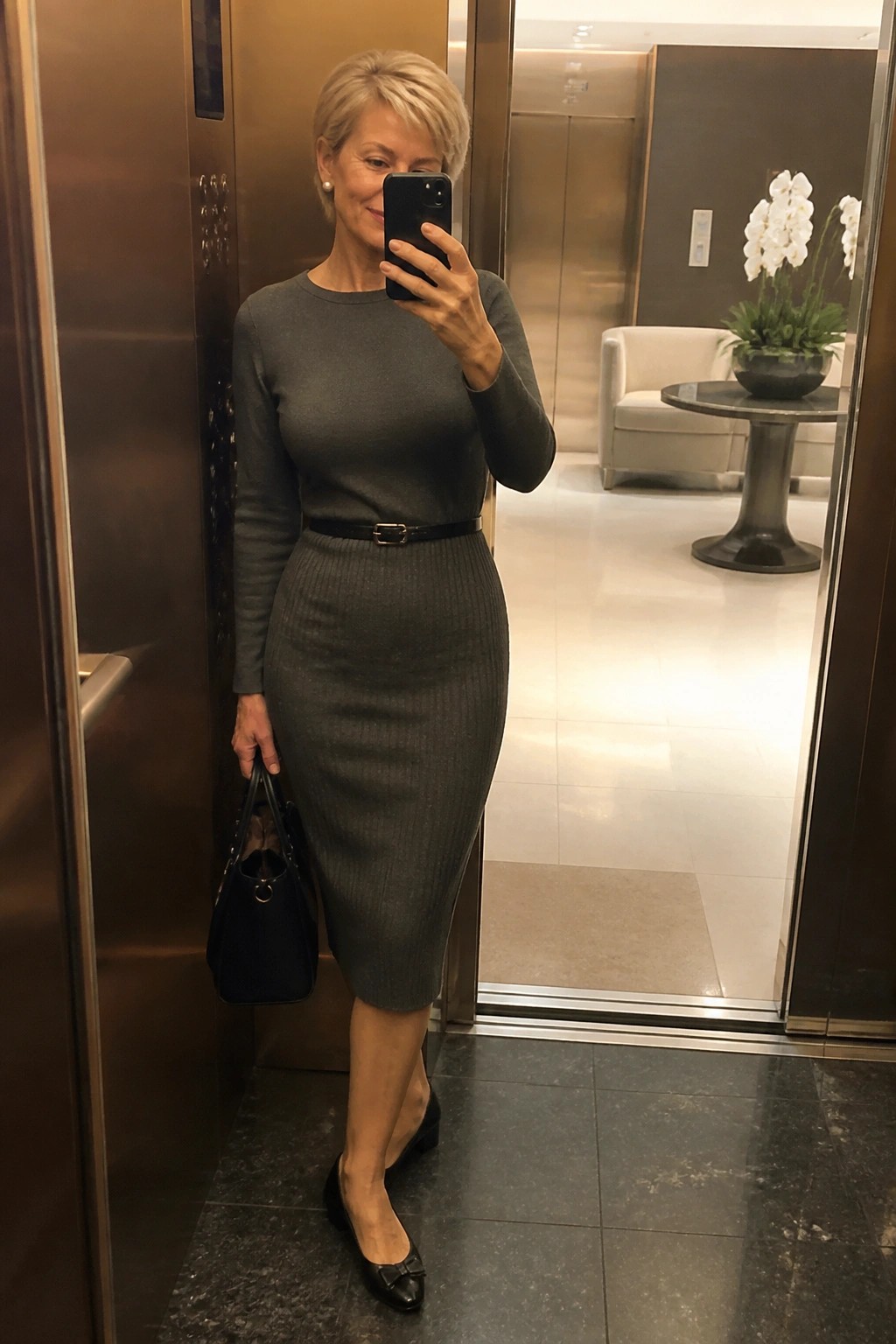 Woman in gray midi dress taking mirror selfie in modern lobby with orchids