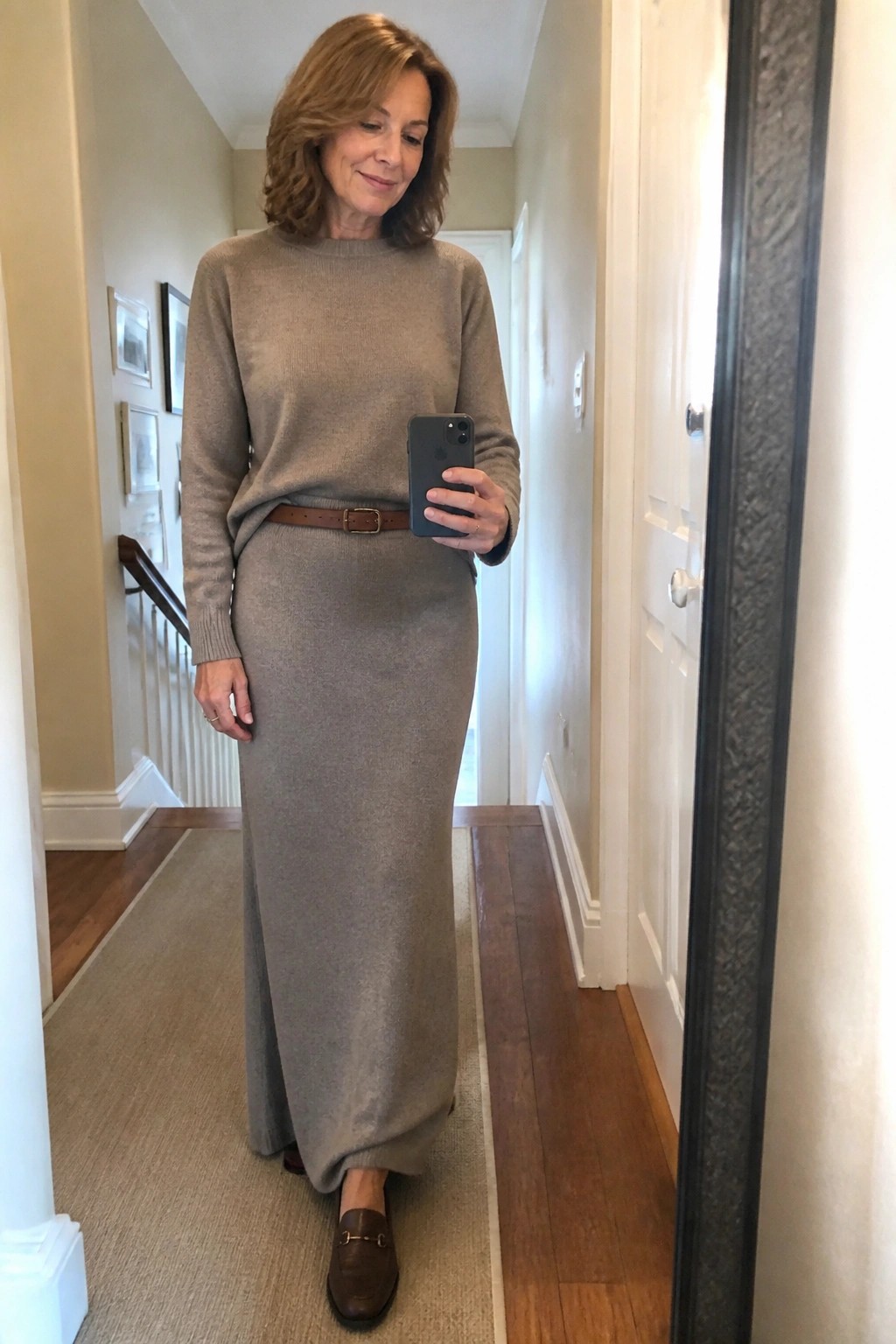 Woman in beige sweater and long skirt taking mirror selfie in hallway
