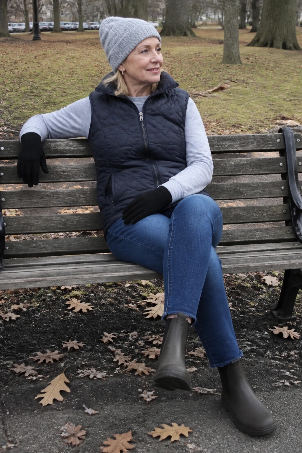 Woman in gray beanie and navy vest seated on leaf-strewn park bench