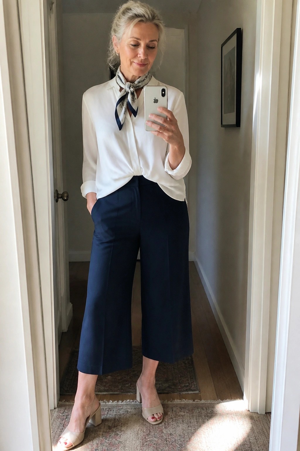 Woman in white blouse with scarf and navy wide-leg pants taking mirror selfie in hallway