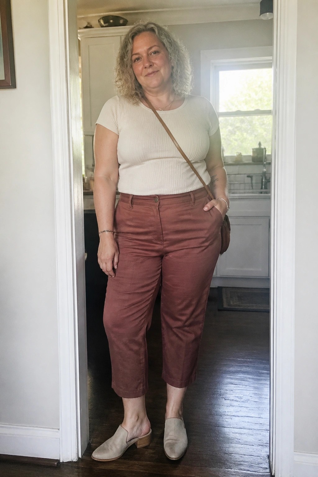 Woman with curly hair in cream top and rust pants standing in kitchen doorway