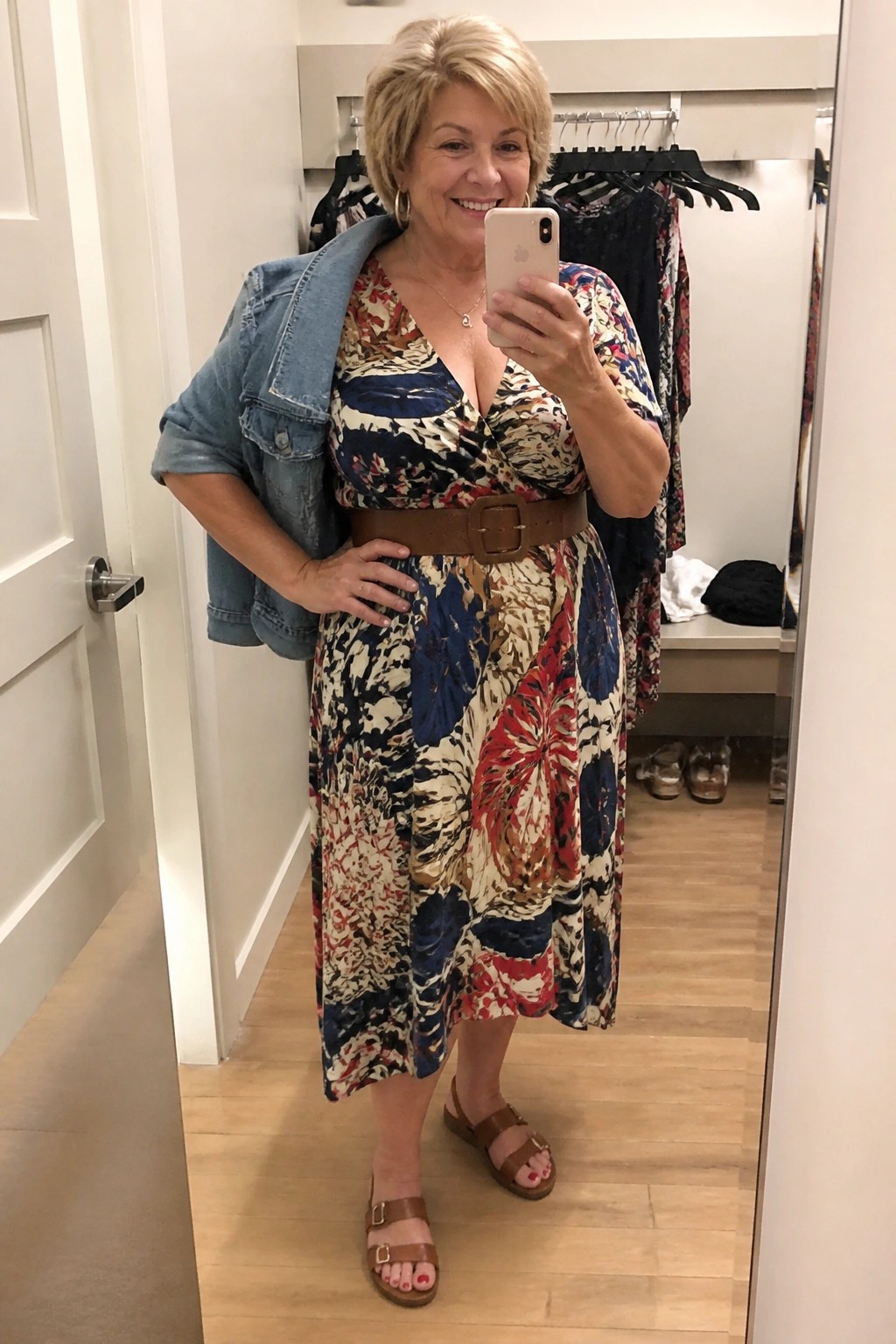 Smiling blonde woman in patterned dress with brown belt and denim jacket in closet