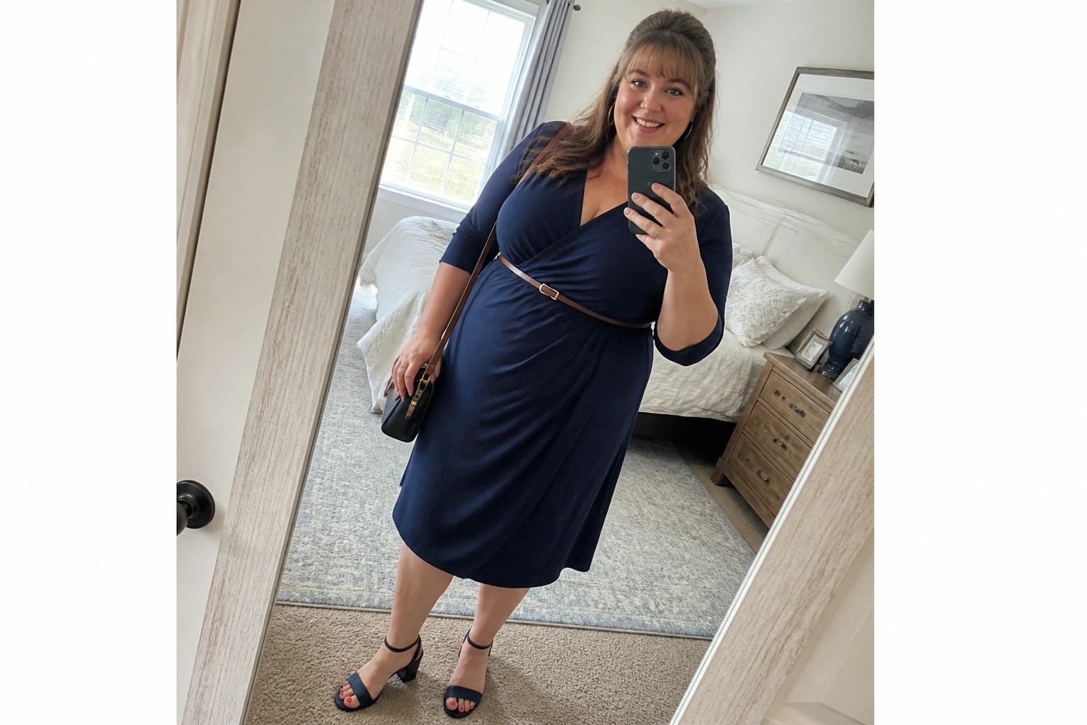 Woman in navy wrap dress taking mirror selfie in bedroom with handbag