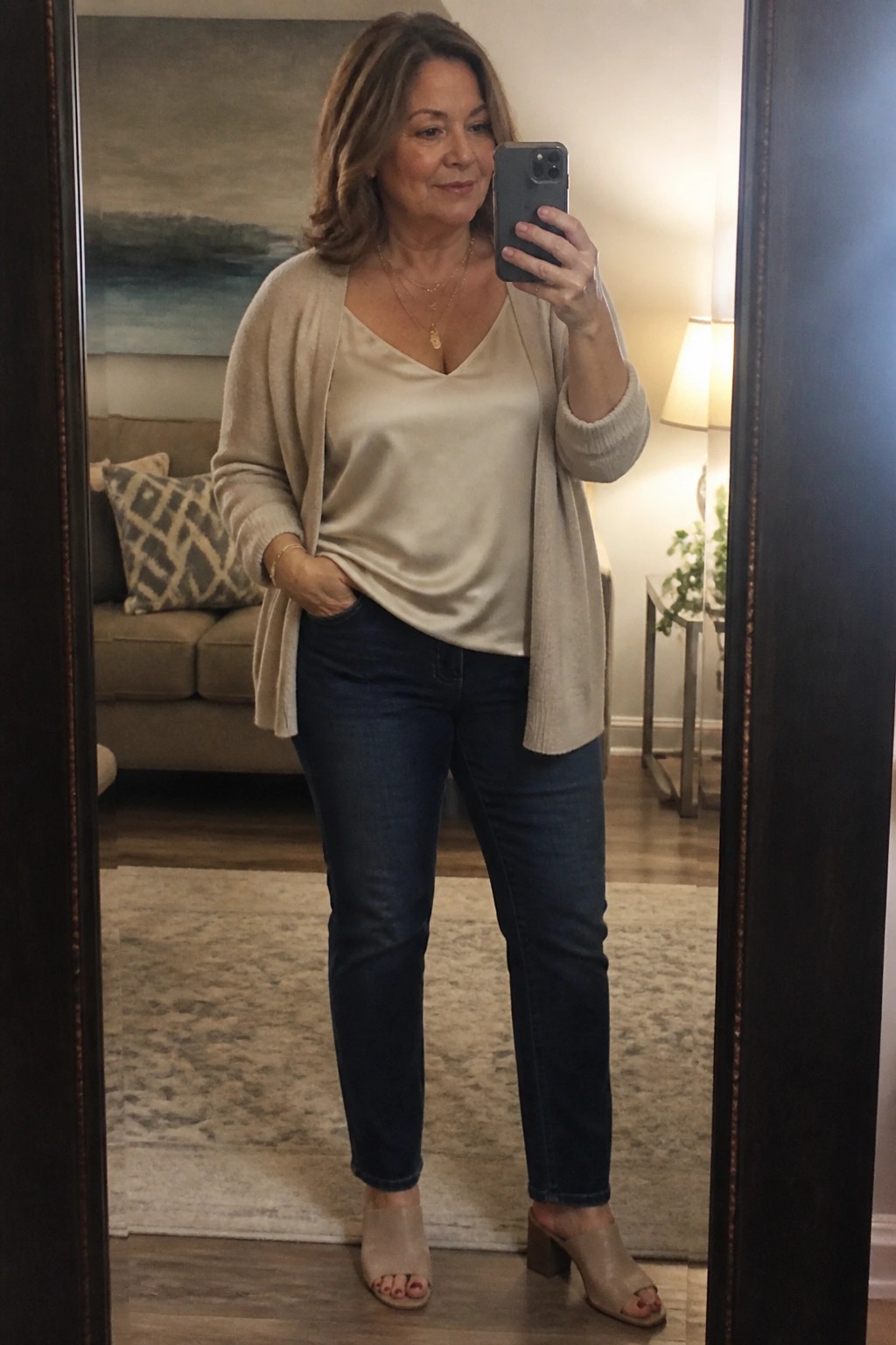 A plus-size woman over 50 in a beige open cardigan layered over a matching V-neck top, dark blue jeans, and beige block heel mules, holding a phone for a mirror selfie with gold necklaces visible