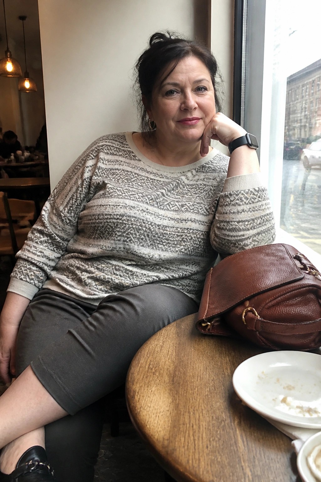 Woman in patterned sweater seated at wooden cafe table with brown leather bag by window