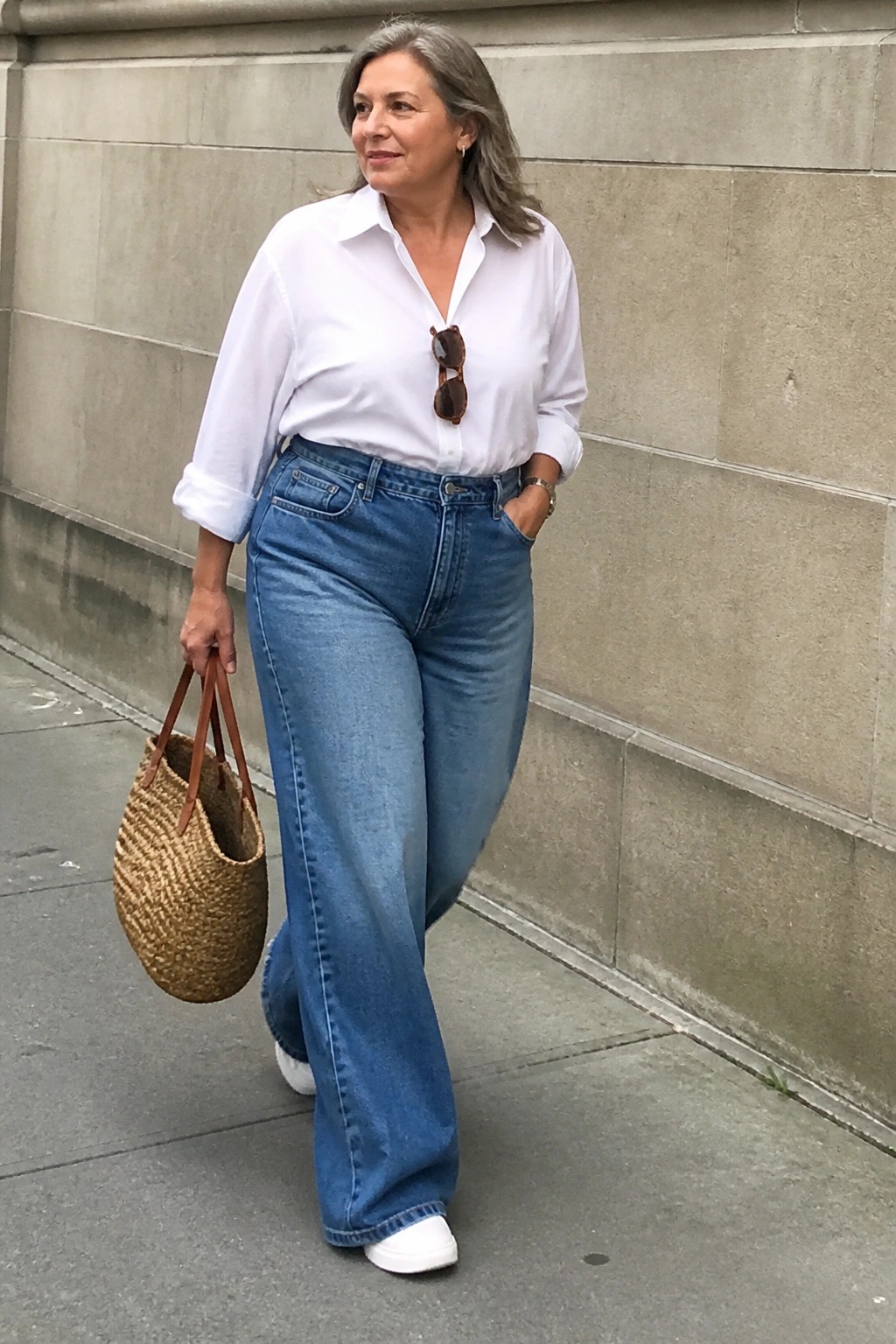 Woman with gray hair in white shirt and blue wide-leg jeans holding woven bag