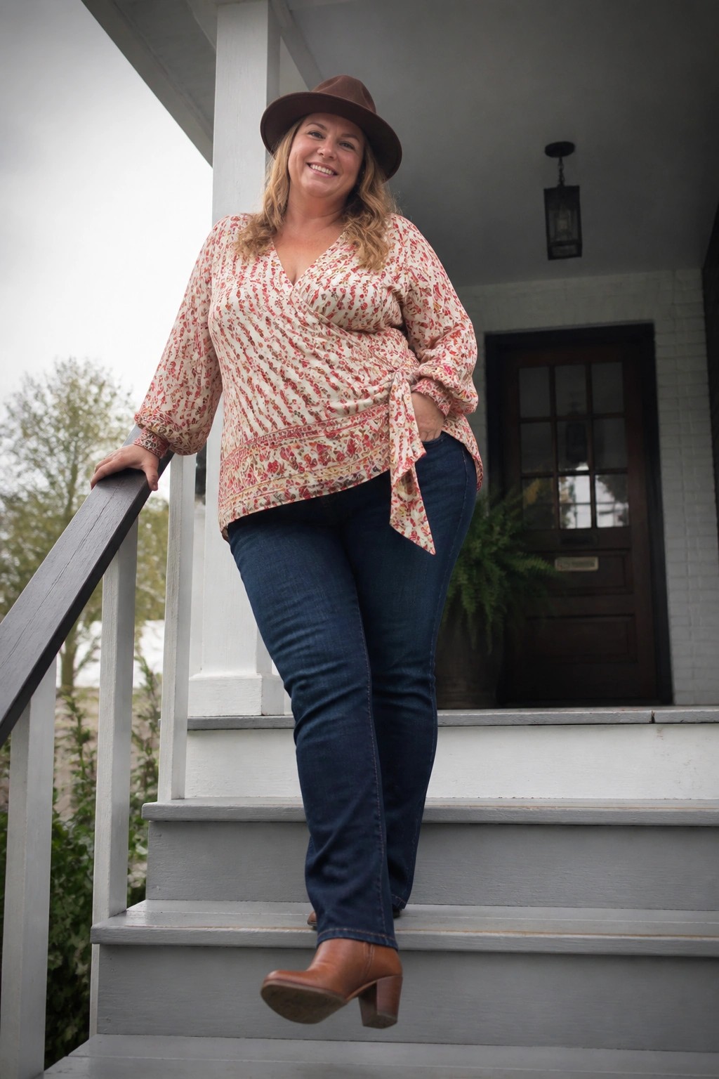 Smiling woman in floral wrap blouse, hat, and jeans on porch steps