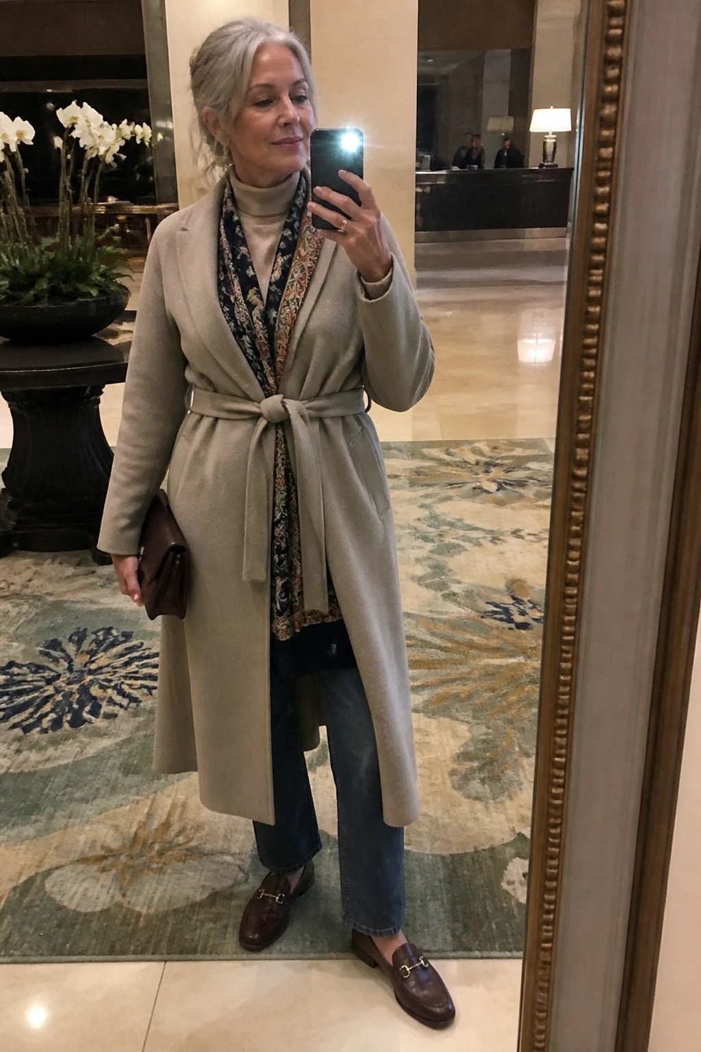 Silver-haired woman in beige coat and jeans taking mirror selfie in hotel lobby.