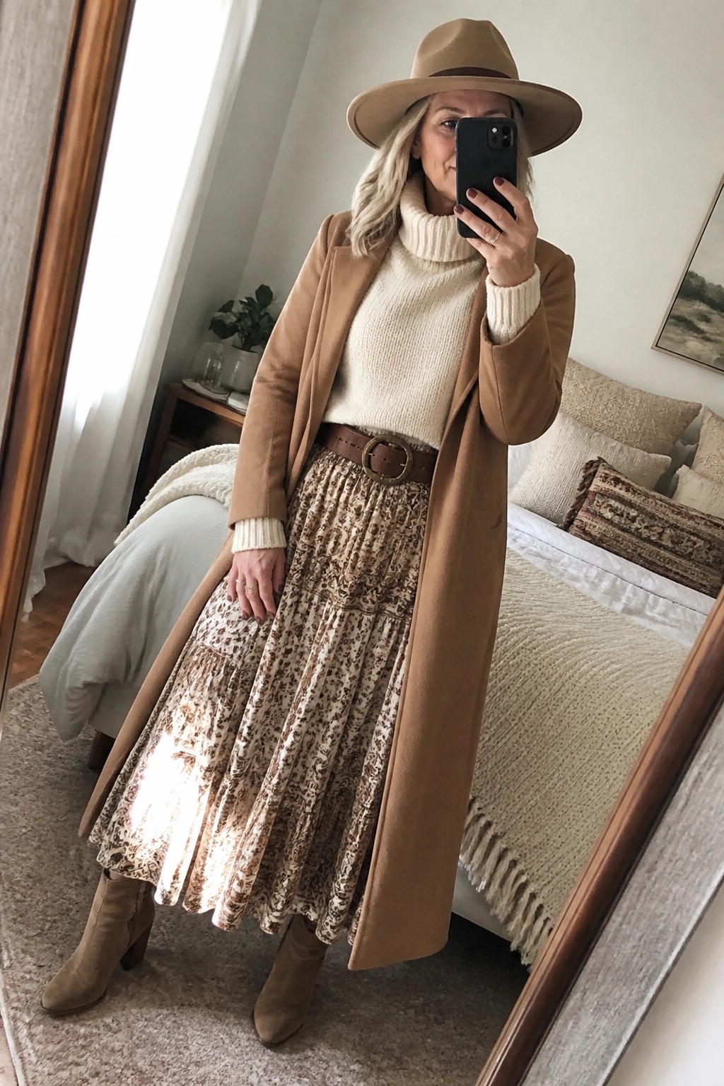 Woman in tan coat, cream turtleneck, patterned skirt, and hat taking mirror selfie