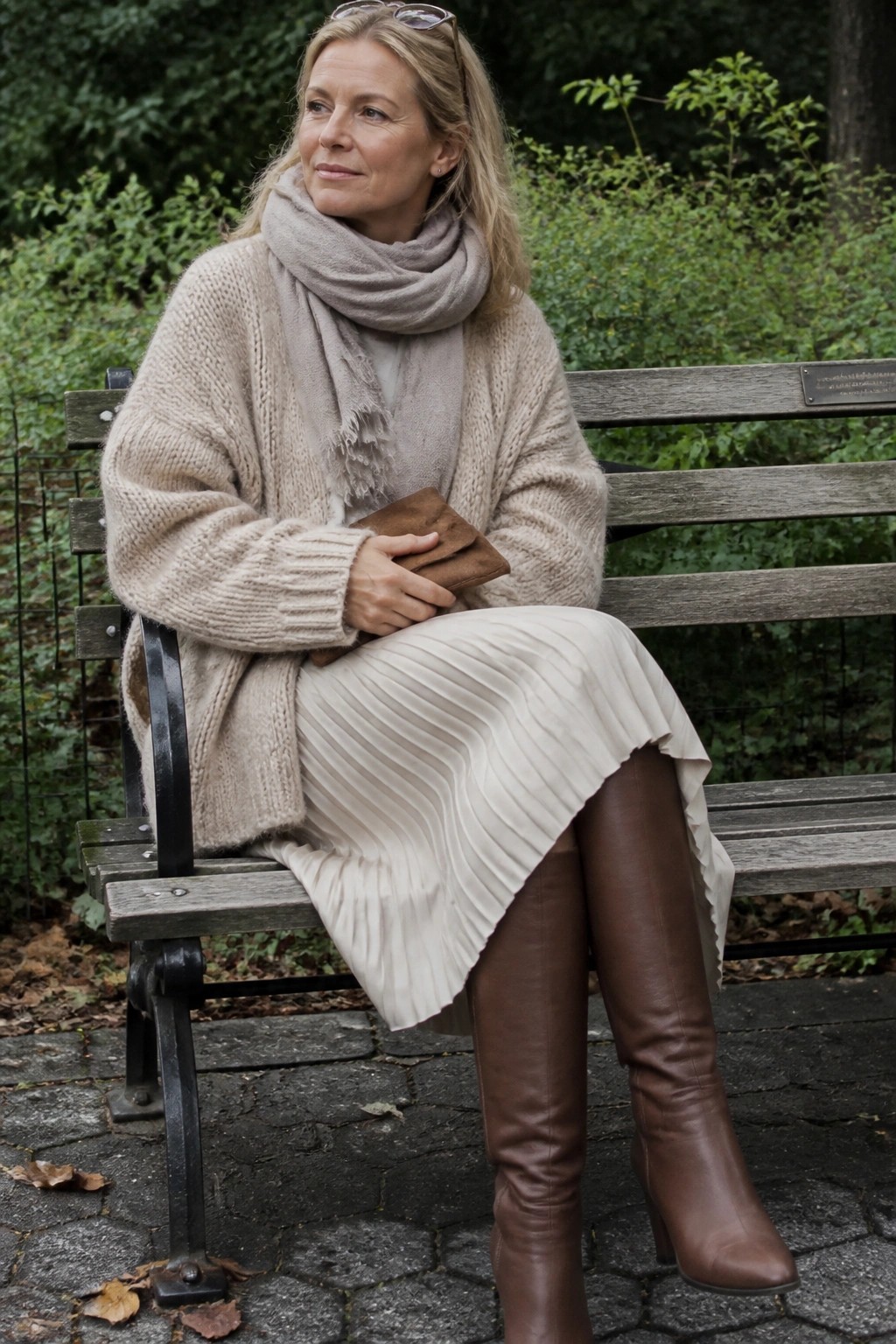 Blonde woman in beige knit sweater, pleated skirt, and brown boots on park bench.