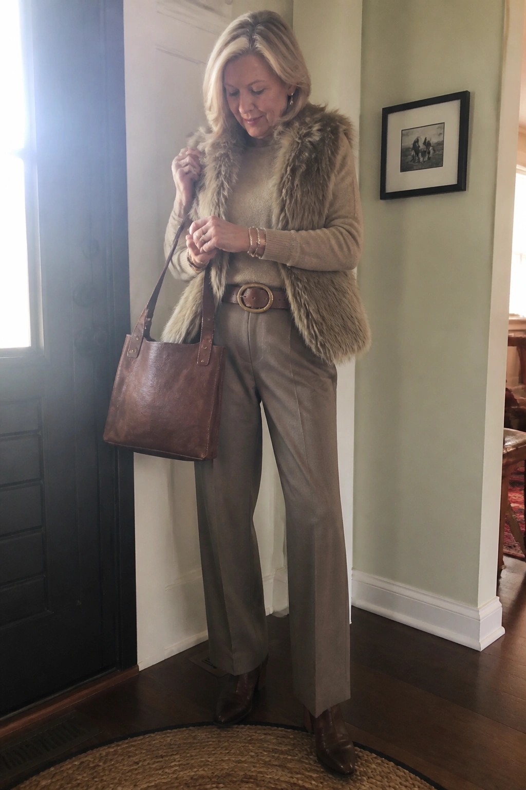 Blonde woman in beige sweater and fur vest with brown leather tote bag indoors.
