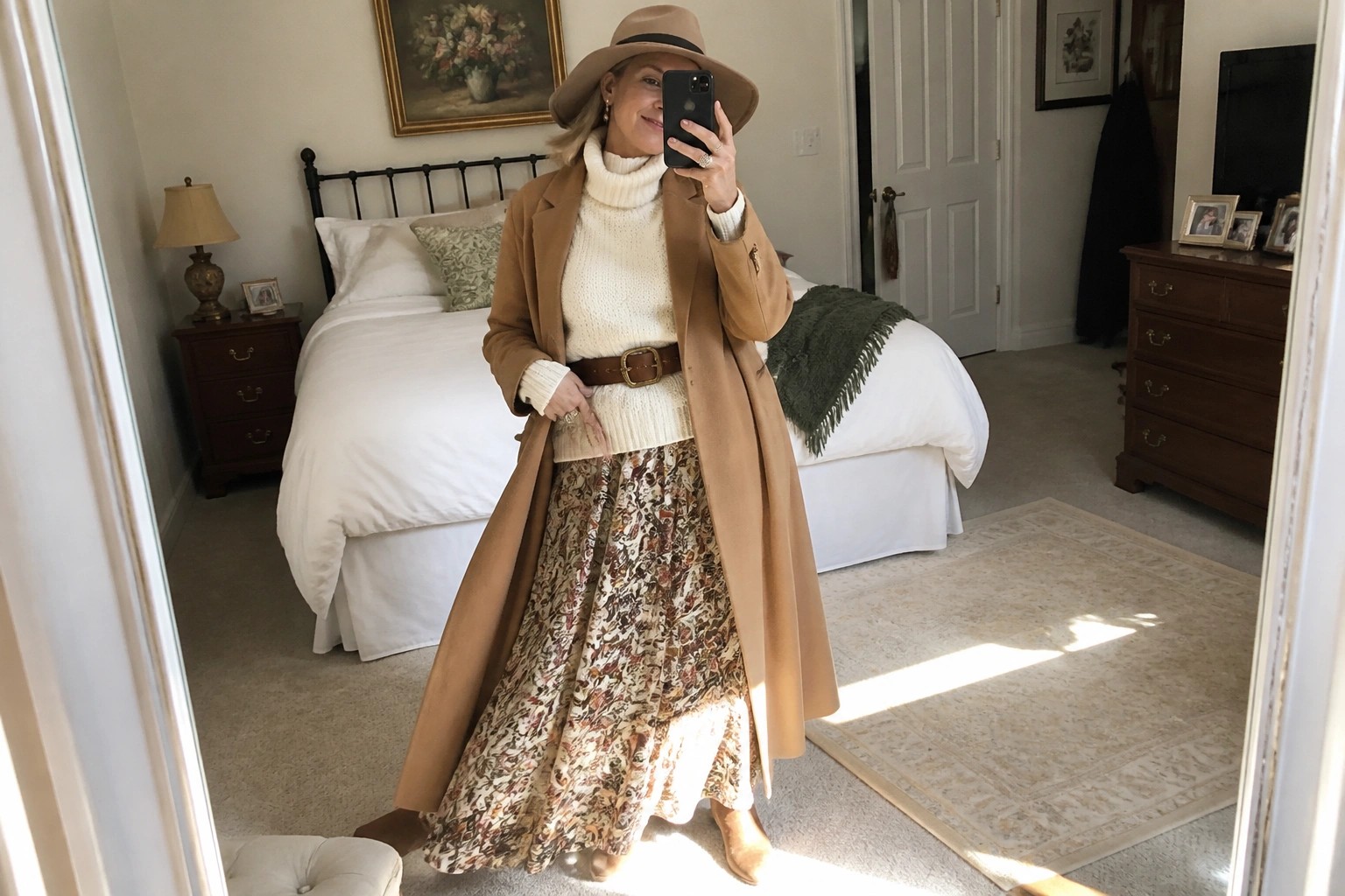 Woman in tan coat, cream turtleneck, patterned skirt, and hat taking mirror selfie