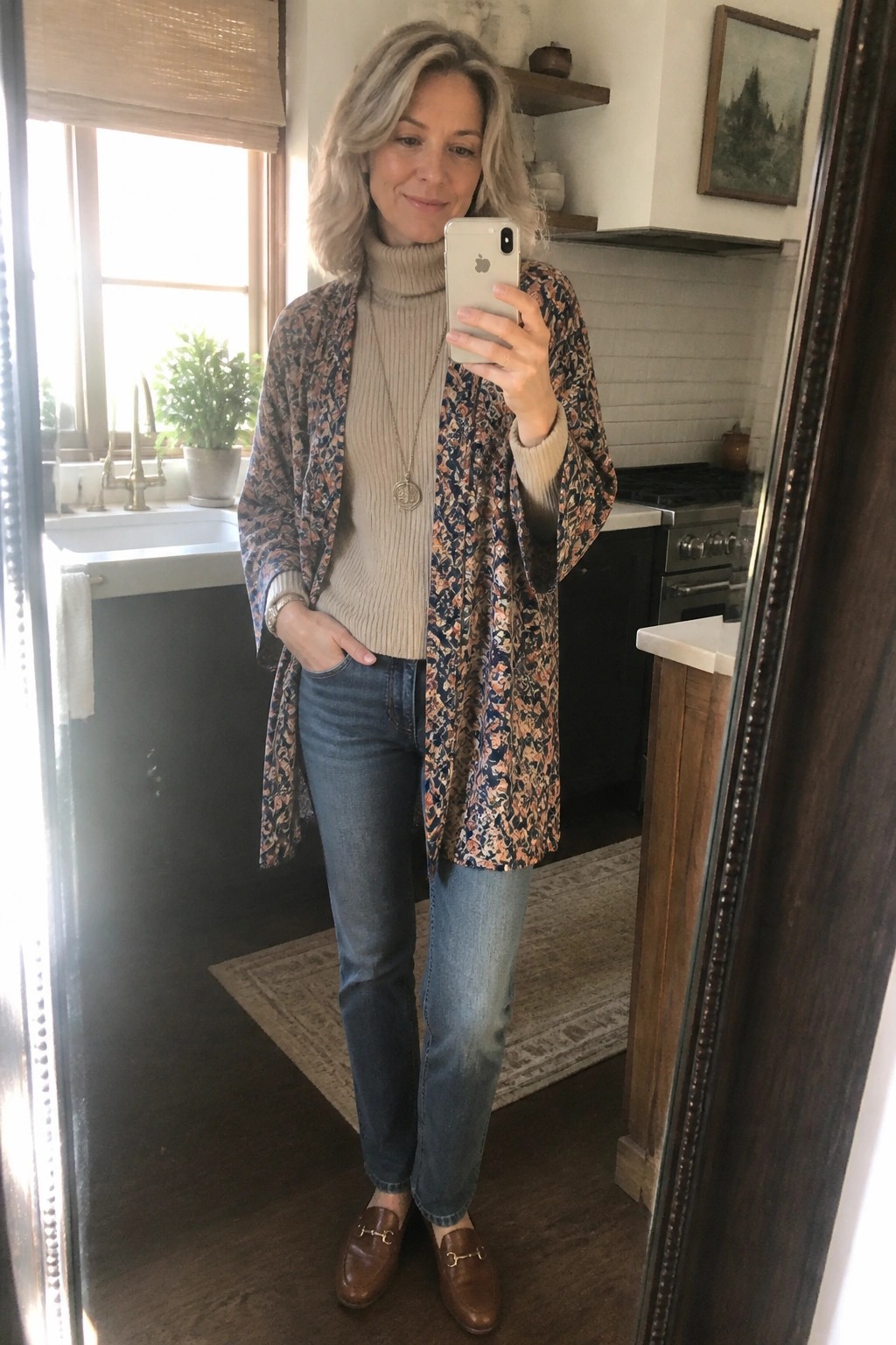 Woman in floral kimono over beige turtleneck taking kitchen mirror selfie.
