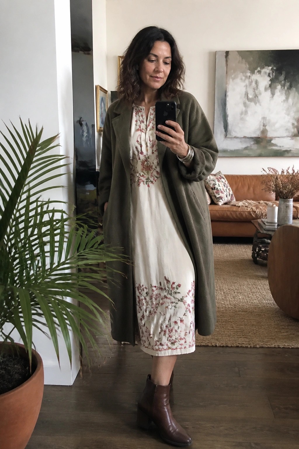 Woman over 50 wearing an olive green wool coat layered over a cream midi dress with pink and green floral embroidery, paired with brown leather ankle boots and silver bracelets, taking a mirror selfie indoors.
