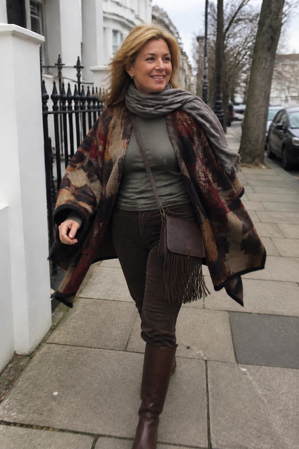 Woman over 50 in layered winter boho outfit featuring a multicolored patterned shawl over an olive top, brown corduroy pants, gray scarf, fringe leather crossbody bag, and knee high brown boots.