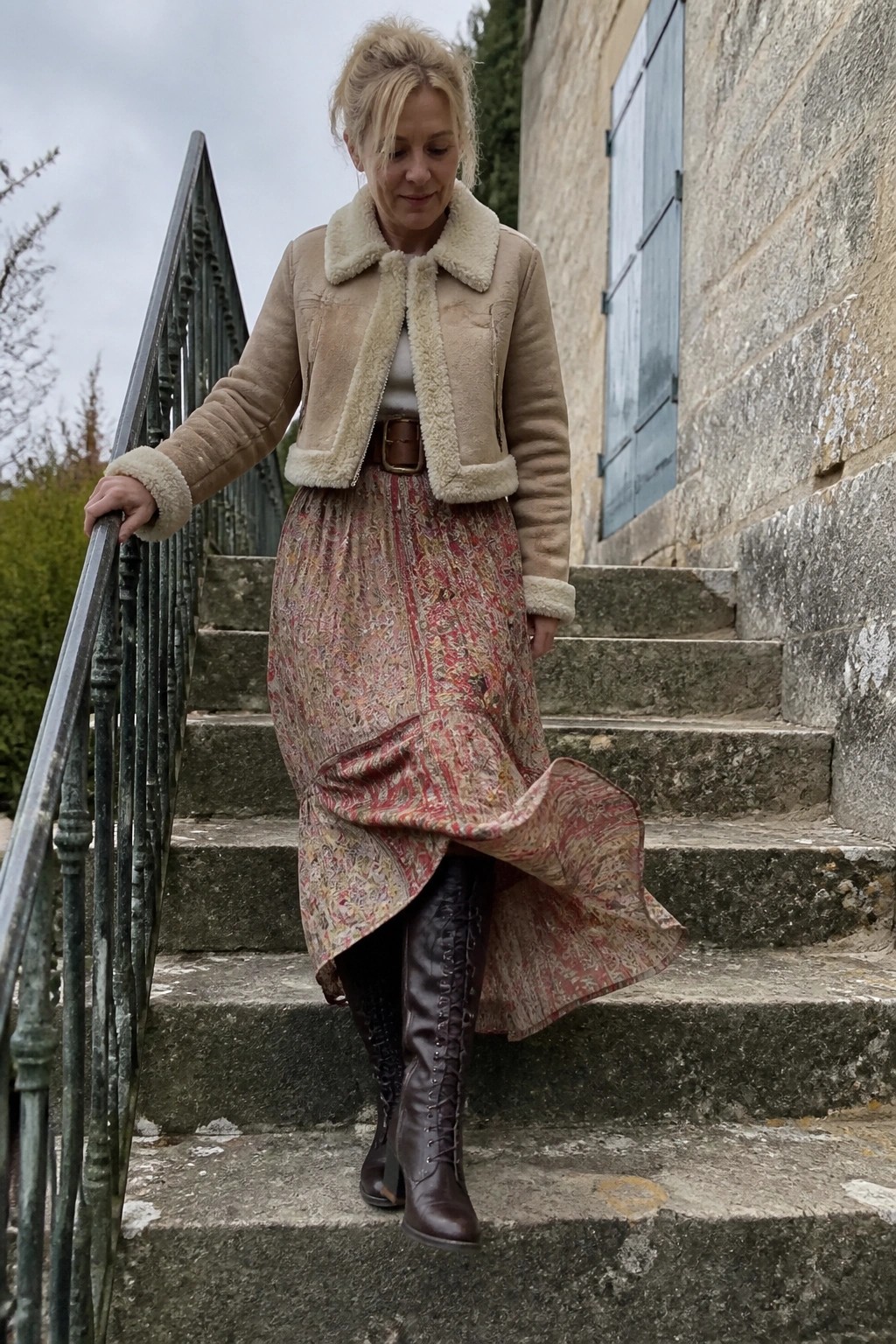Blonde woman in beige shearling jacket and red patterned skirt on stone steps