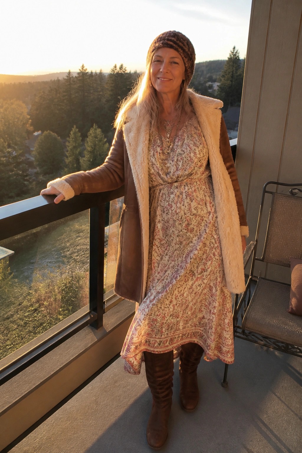 Woman in floral dress and shearling jacket on balcony overlooking forest at sunset