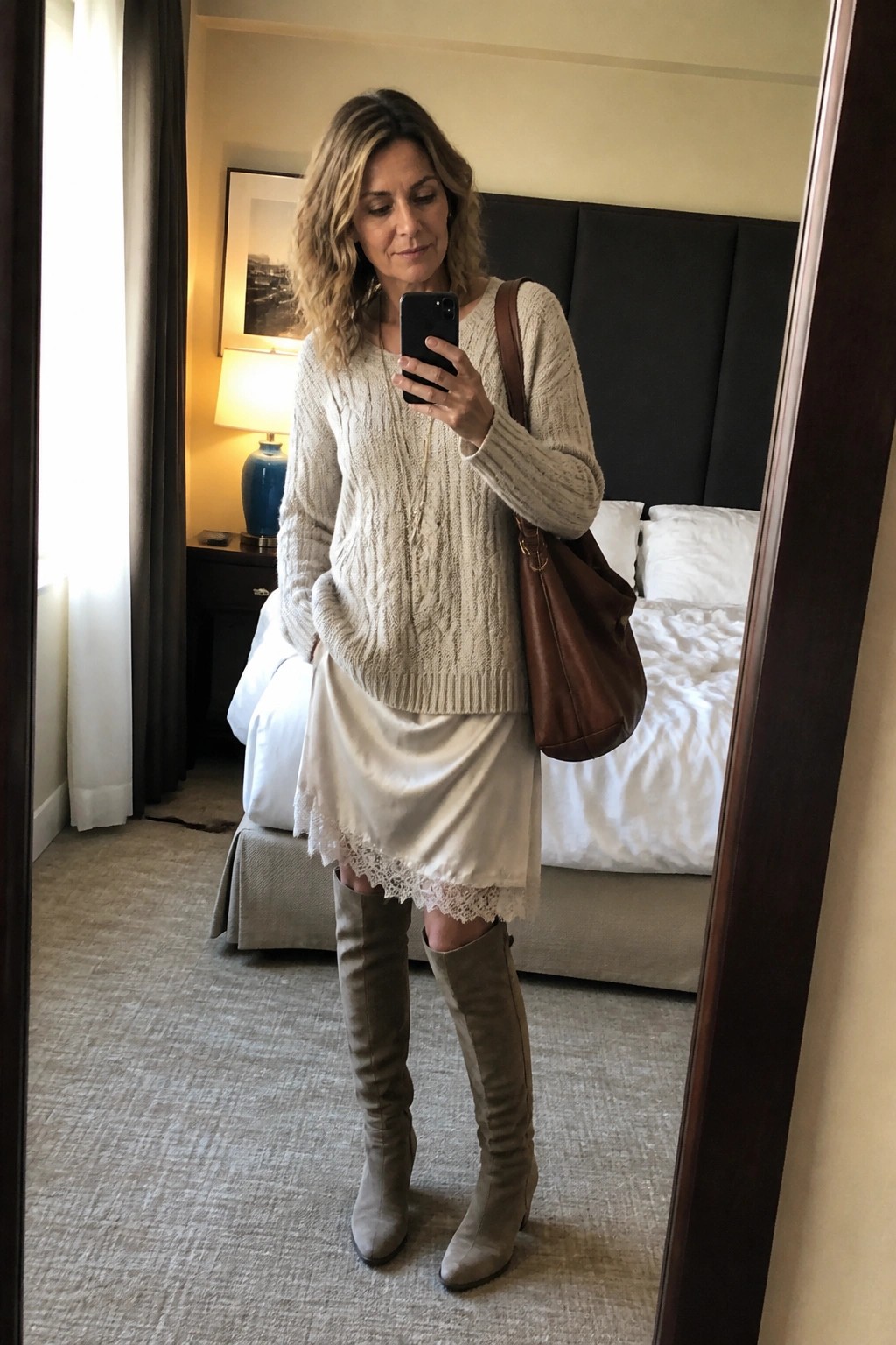 Woman in cream cable-knit sweater and white lace slip dress taking hotel mirror selfie