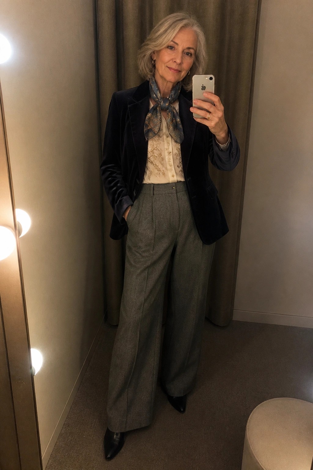 Silver-haired woman in navy velvet blazer and gray trousers taking mirror selfie.