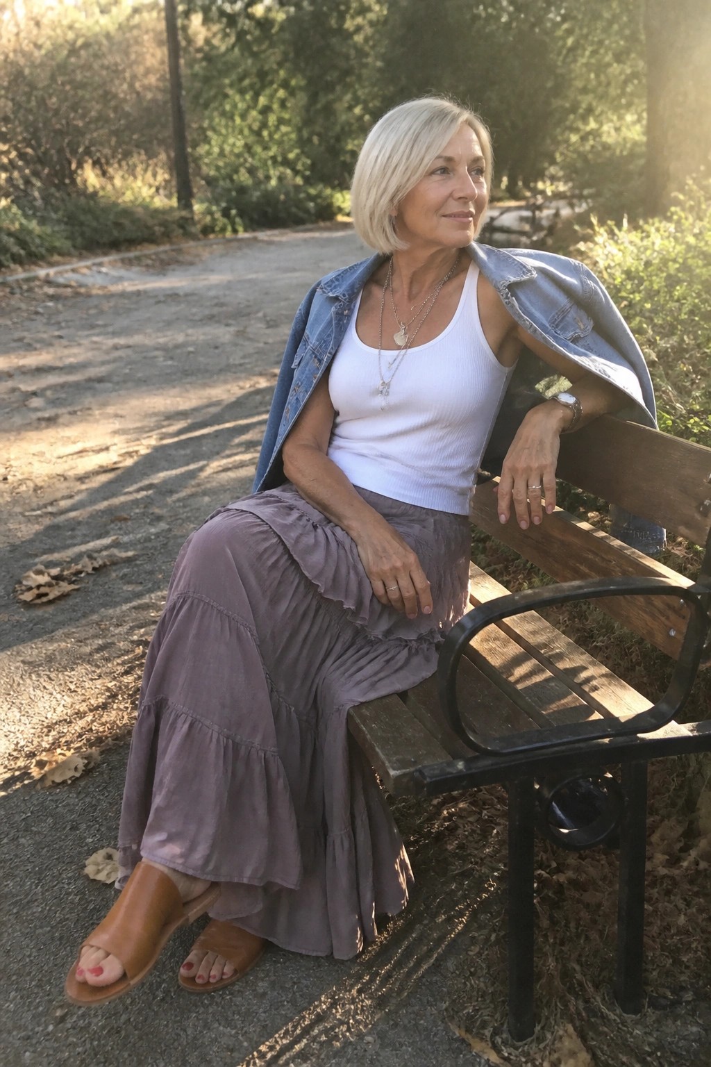 Blonde woman in white tank top, denim jacket, and tiered purple skirt on park bench.